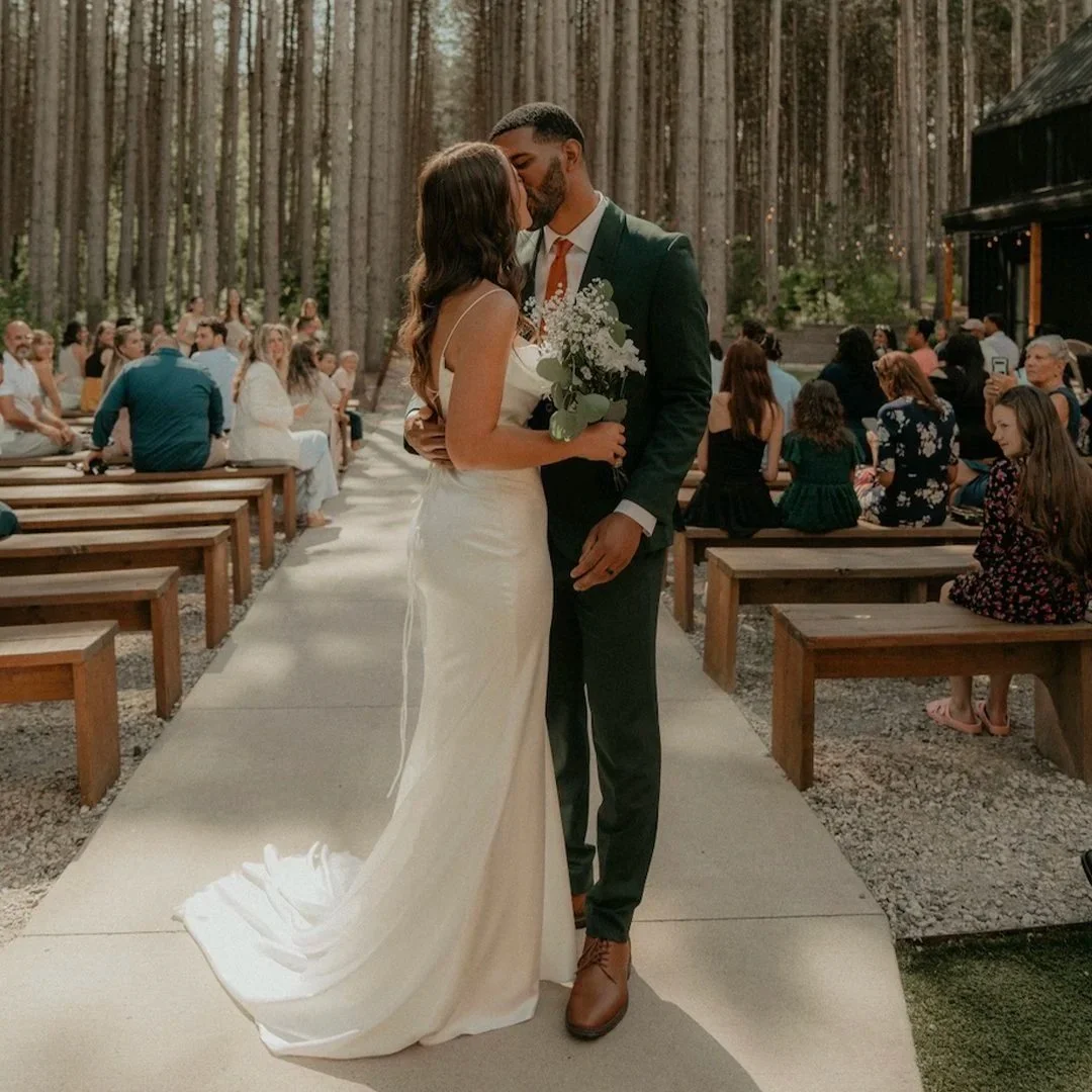A bride and groom kiss at their outdoor Minnesota wedding ceremony in a wooded area, with guests seated on wooden benches and trees in the background.