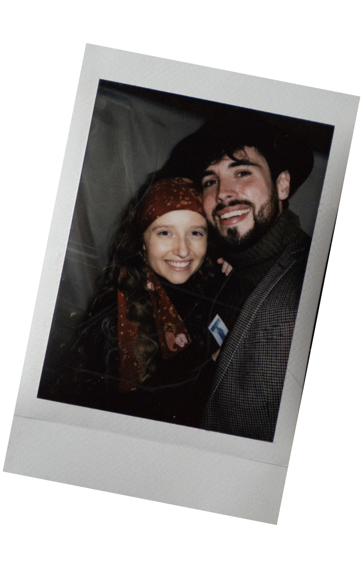 A young woman with long curly hair, wearing a red headscarf decorated with flowers, and a man with dark hair and beard, smiling, dressed in a dark coat and plaid jacket, both smiling and posing for a photo.
