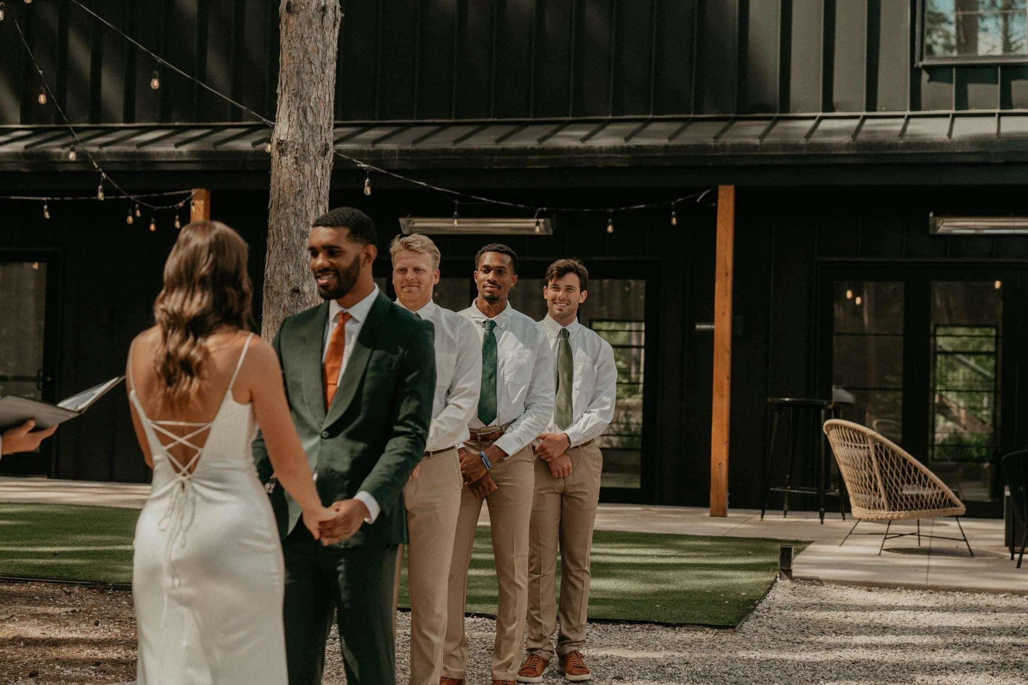 A wedding ceremony outdoors with five men in dress shirts and ties, the bride and groom holding hands, surrounded by natural and modern decor.