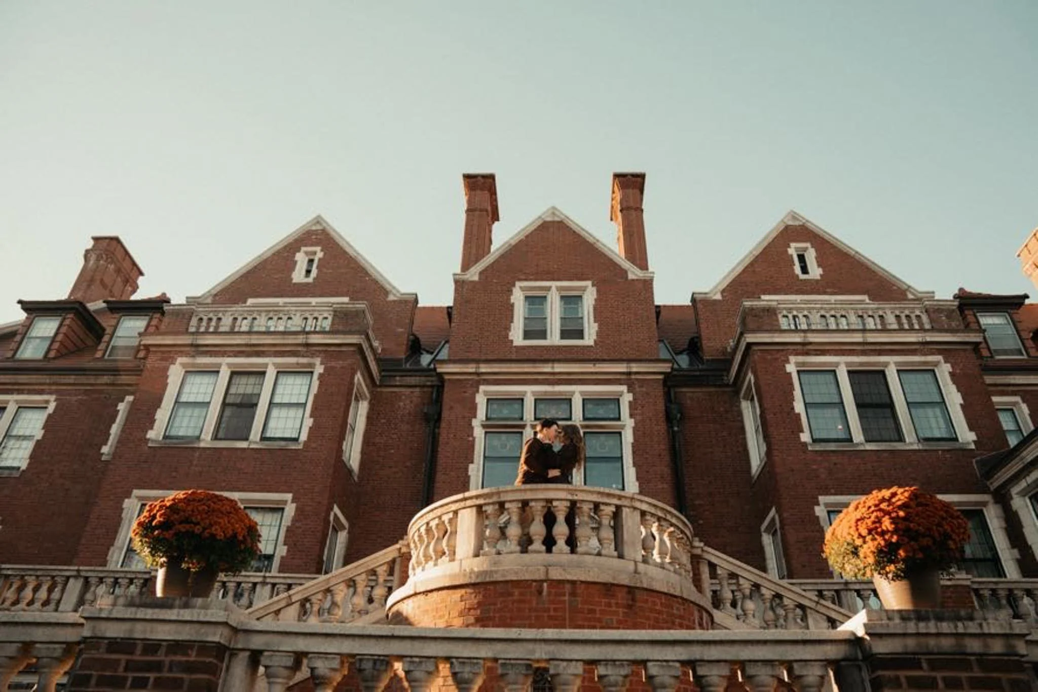 A couple kissing on a curved balcony in front of a large brick mansion with symmetrical windows and a central gabled roof, decorated with flowerpots on the railing.