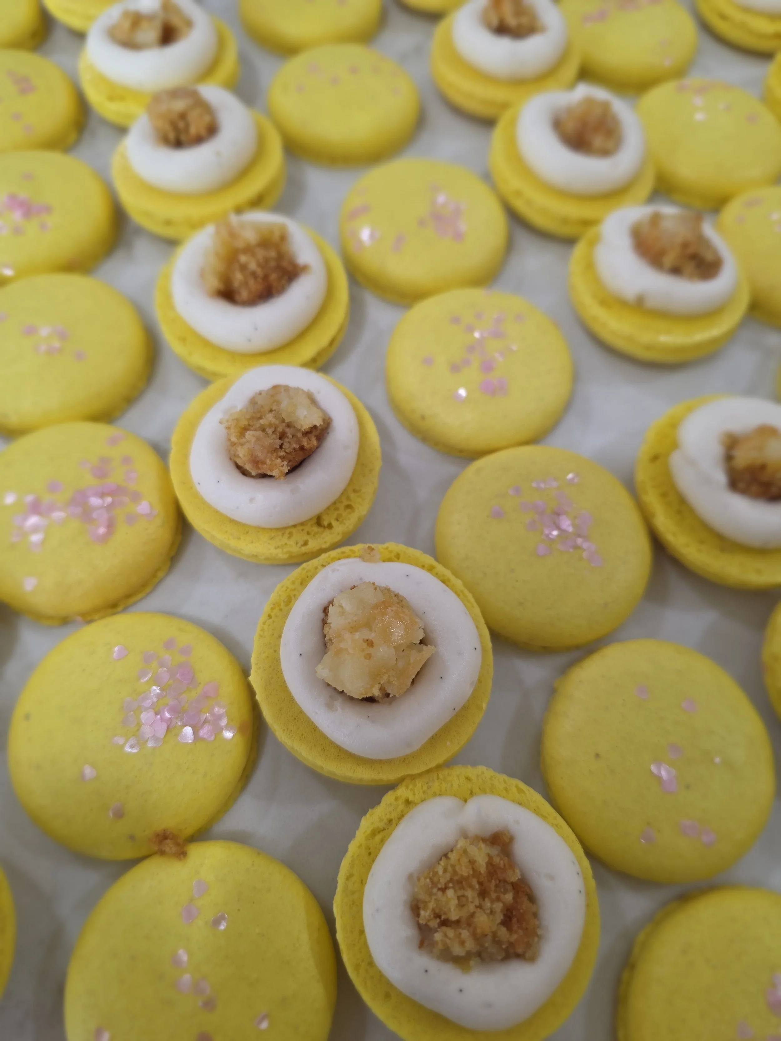 Yellow macarons with pink sprinkles and some topped with white icing and a small crumbly topping inside.