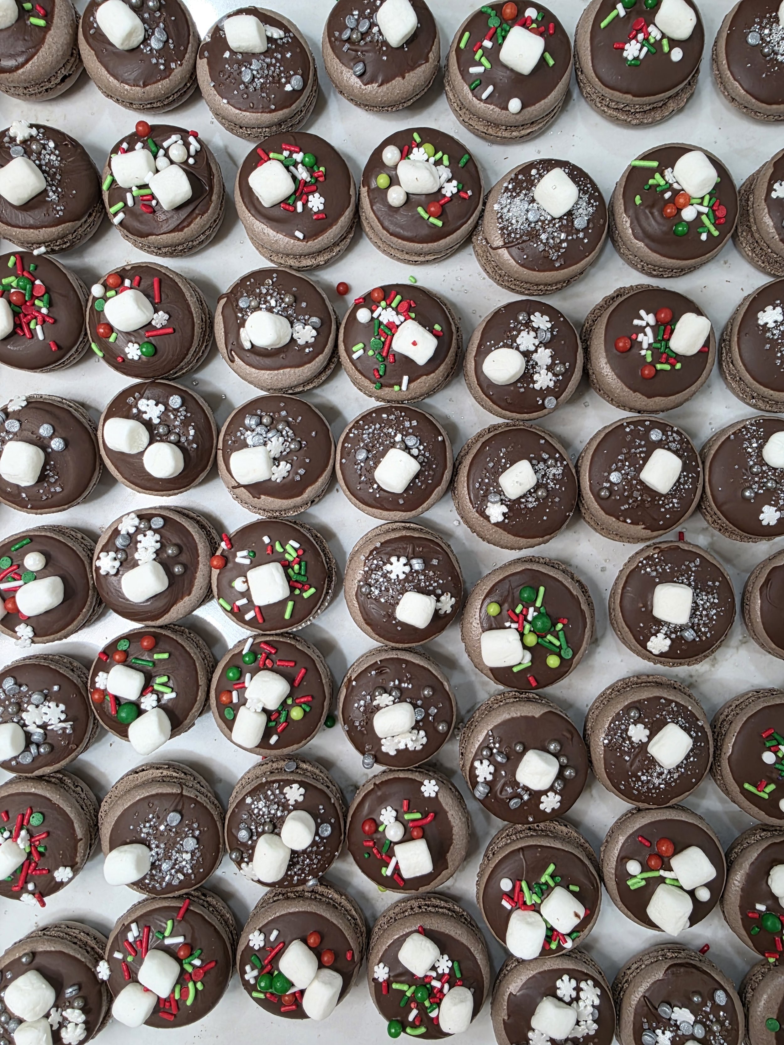 Multiple decorated chocolate macarons with festive sprinkles, mini marshmallows, and sugar snowflake and pearl decorations.