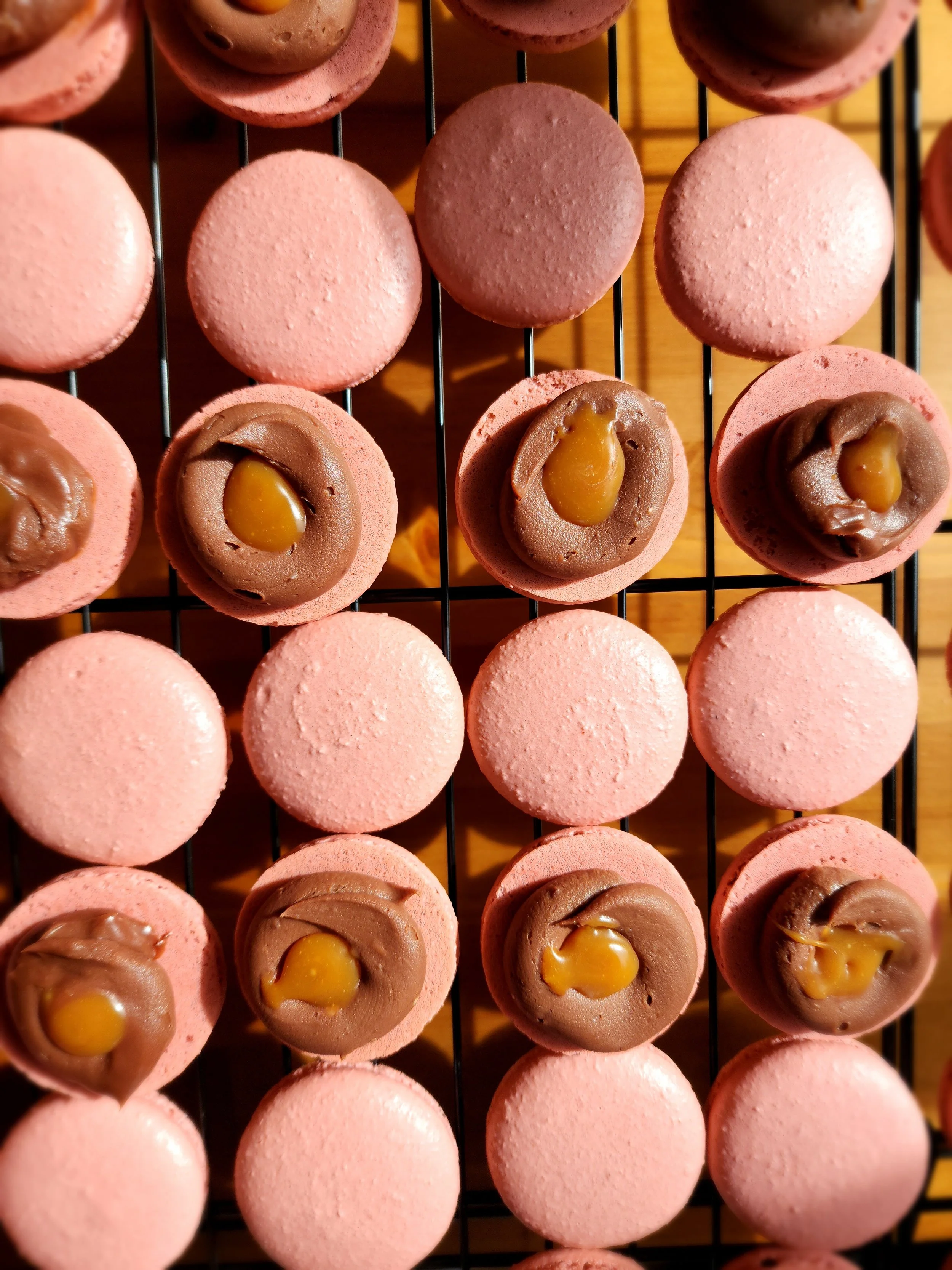 Pink macarons on a wire rack, some with chocolate and caramel filling on top.