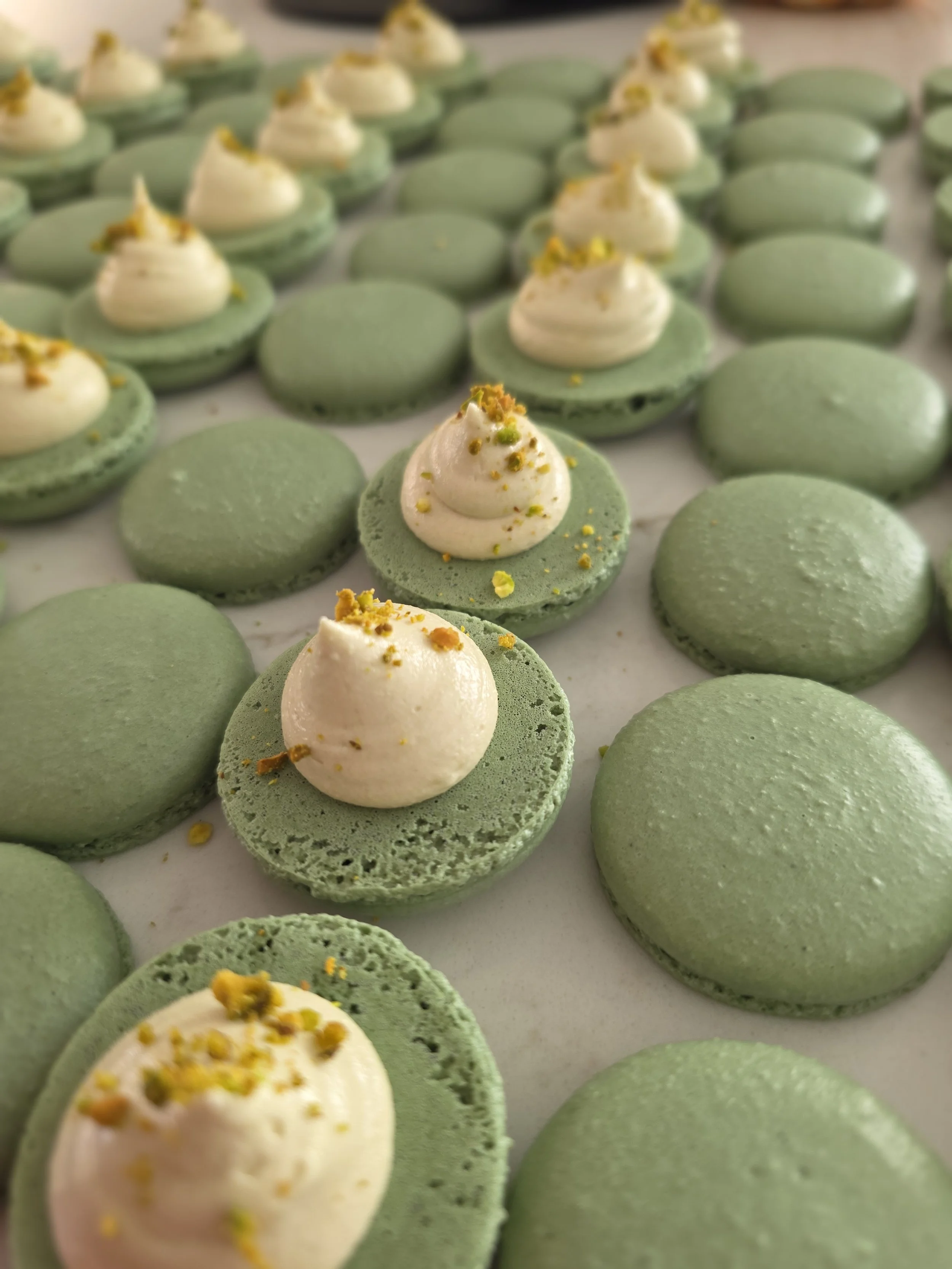 Close-up of green macarons topped with white cream and sprinkled with crushed pistachios.