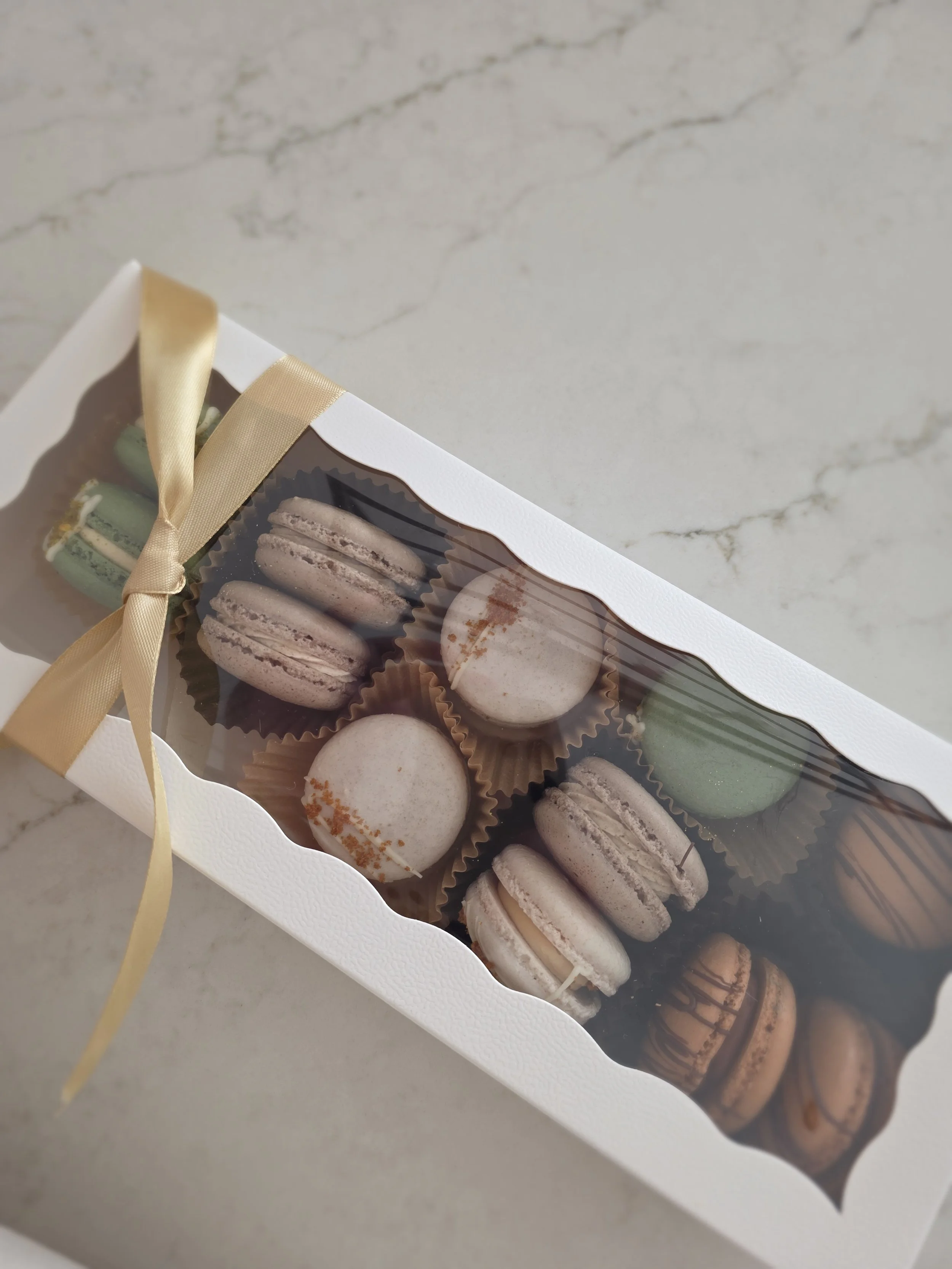 A white gift box with a transparent window and a gold ribbon, containing assorted macarons of various colors and flavors.