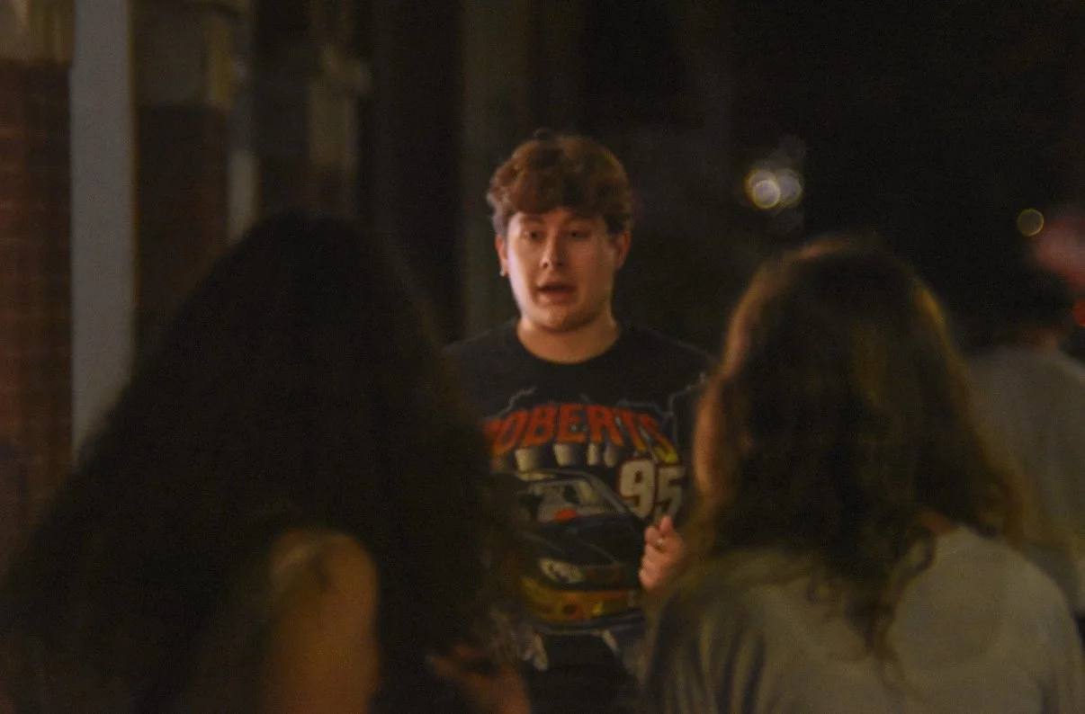 A young man with curly hair in a black t-shirt speaking to two women with long dark hair outdoors at night.