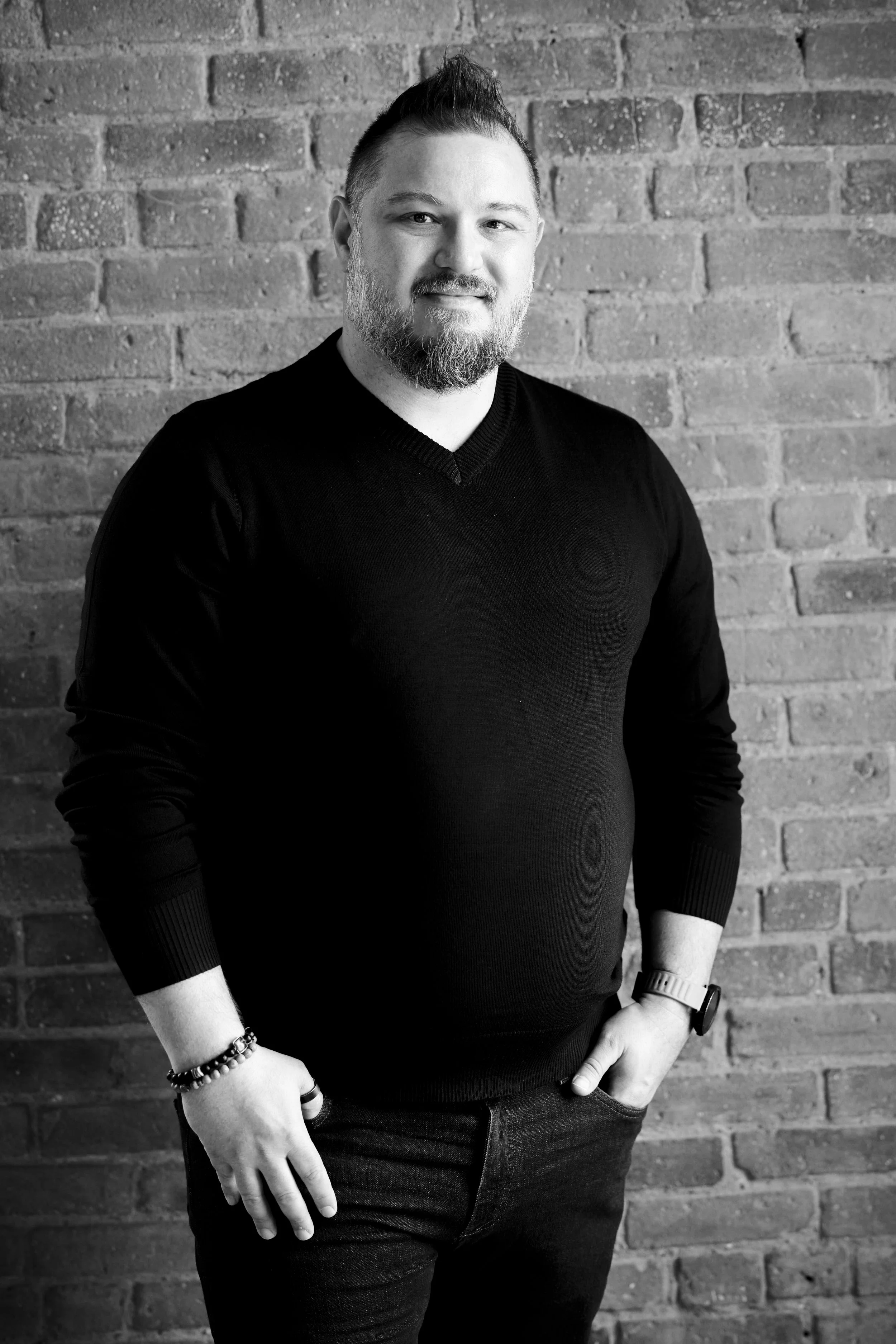 A man with a beard and short hair, wearing a black sweater, standing against a brick wall. He is smiling slightly with one hand in his pocket and wearing a watch and bracelets.