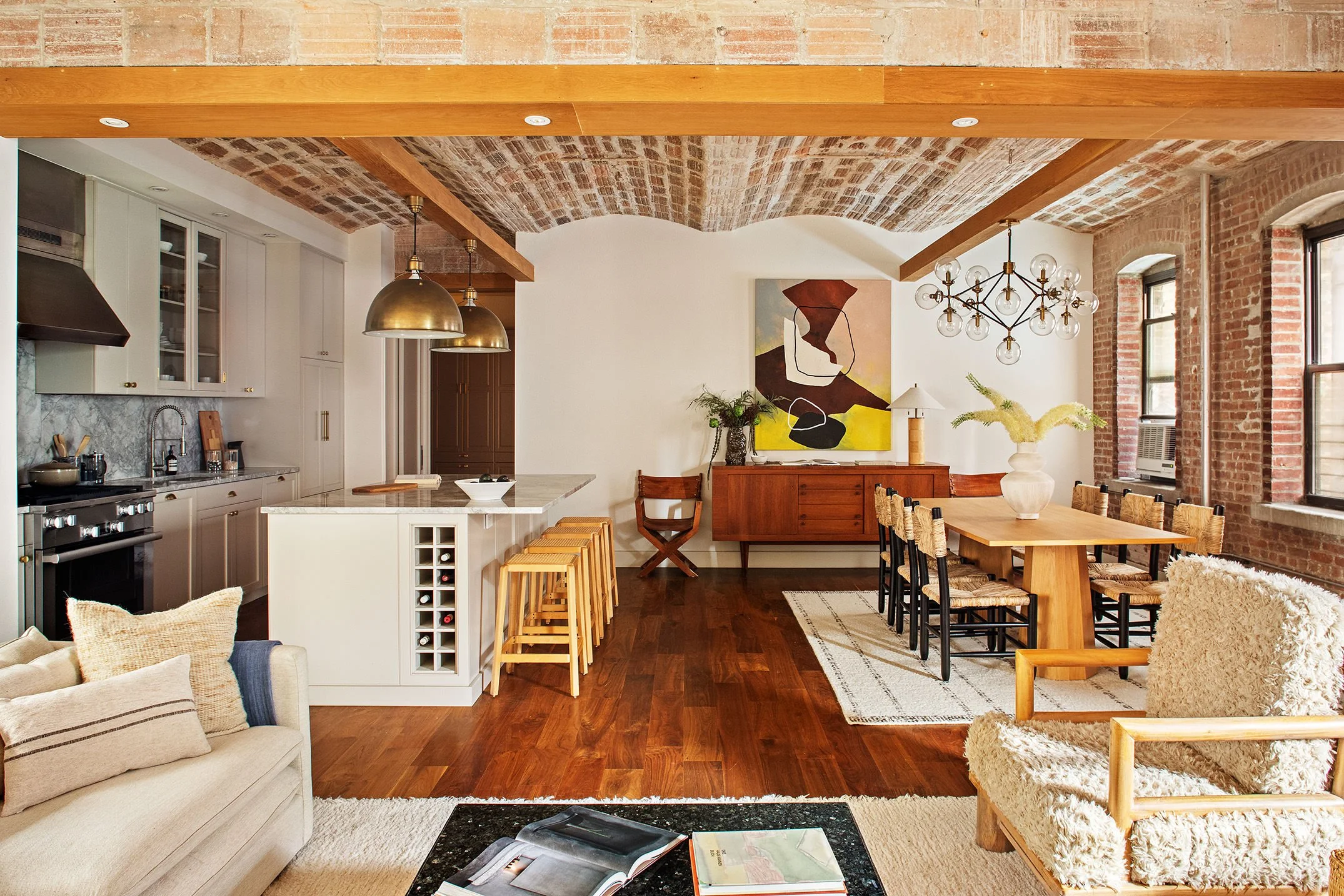  view of kitchen and dining room from living room, on the left is a grey couch and on the right are two comfortable chairs with wooden arm rests 