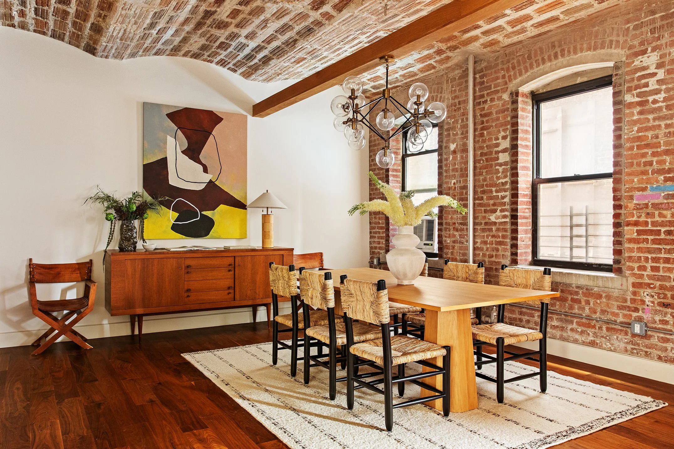  cool toned wood dining table with rattan chairs on a white striped rug over a warm toned floor, a brick accent wall with windows is seen to the right and a warm toned credenza is seen in the back left 