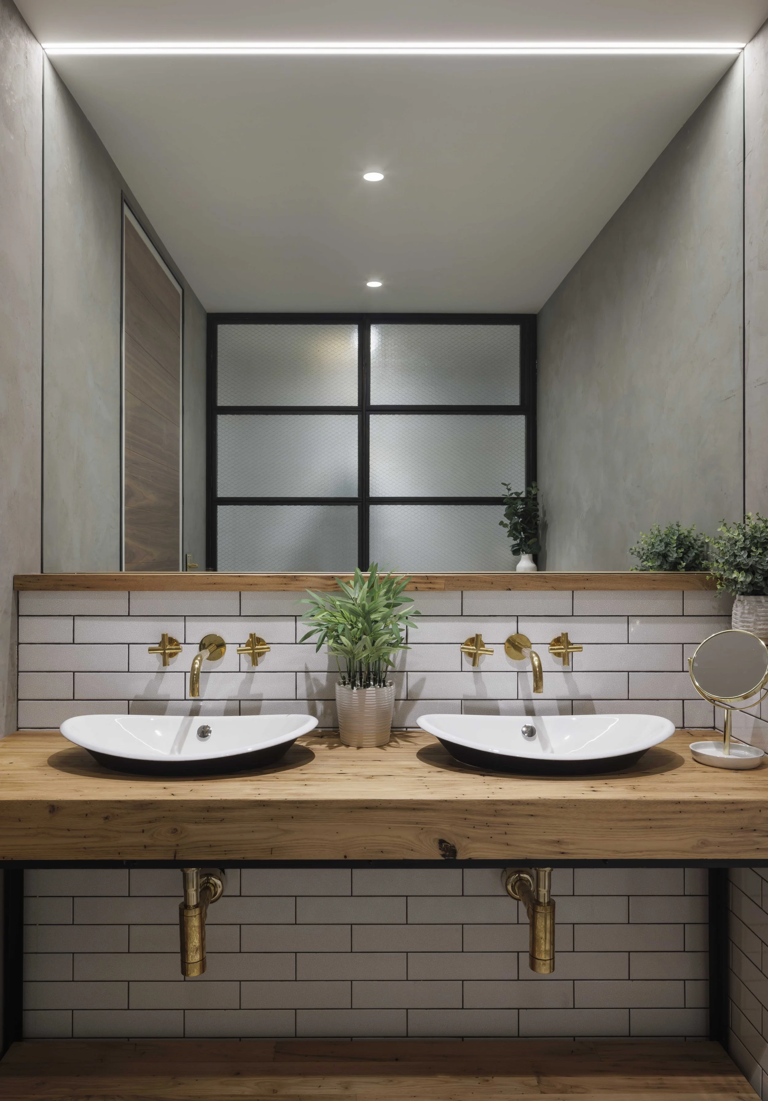  double sinks set in a warm toned wooden counter with a white tiled backsplash and a reflection of window partition walls in the mirror 