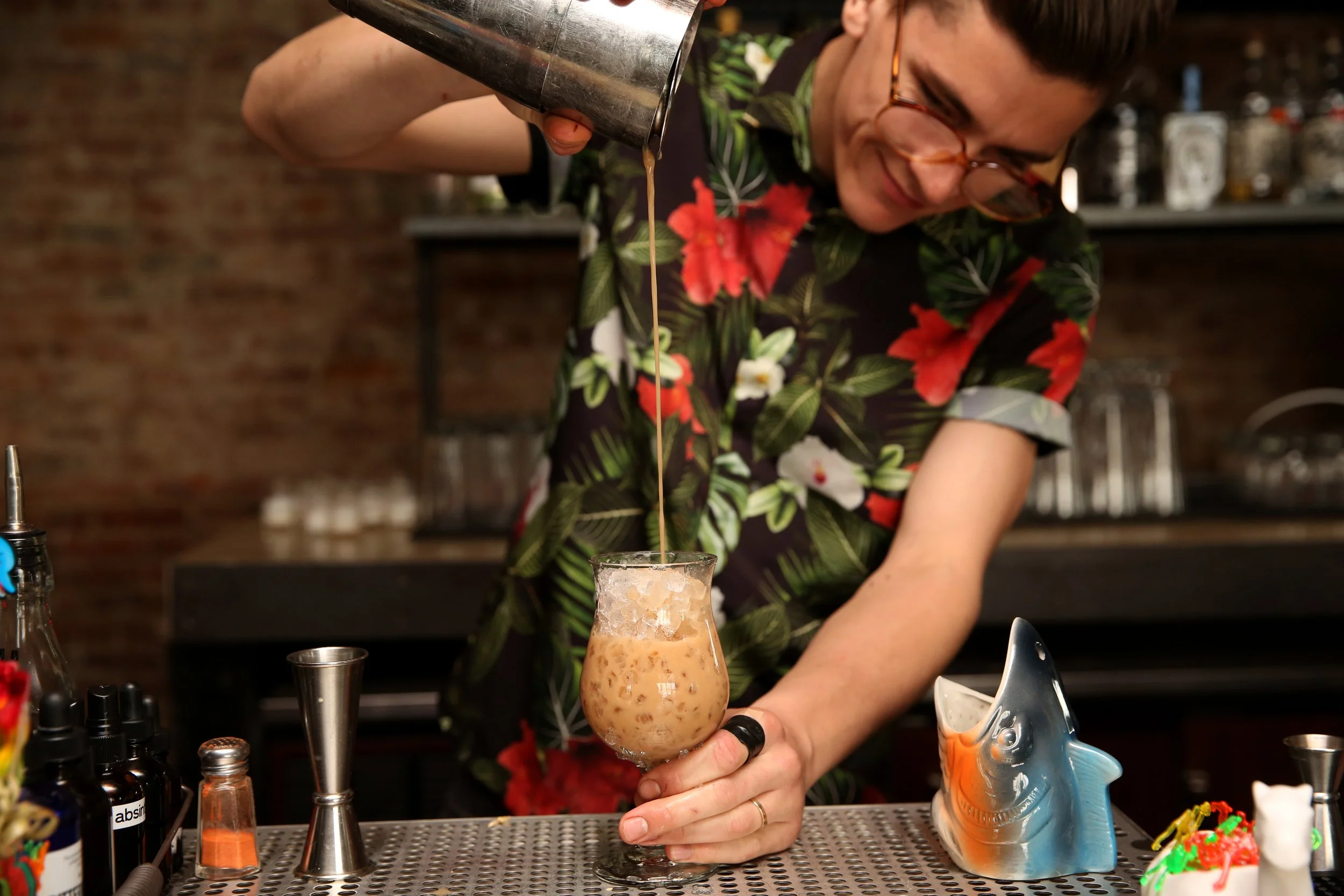  bartender wearing floral button down shirt and glasses pours orange tiki cocktail into cup at bar 