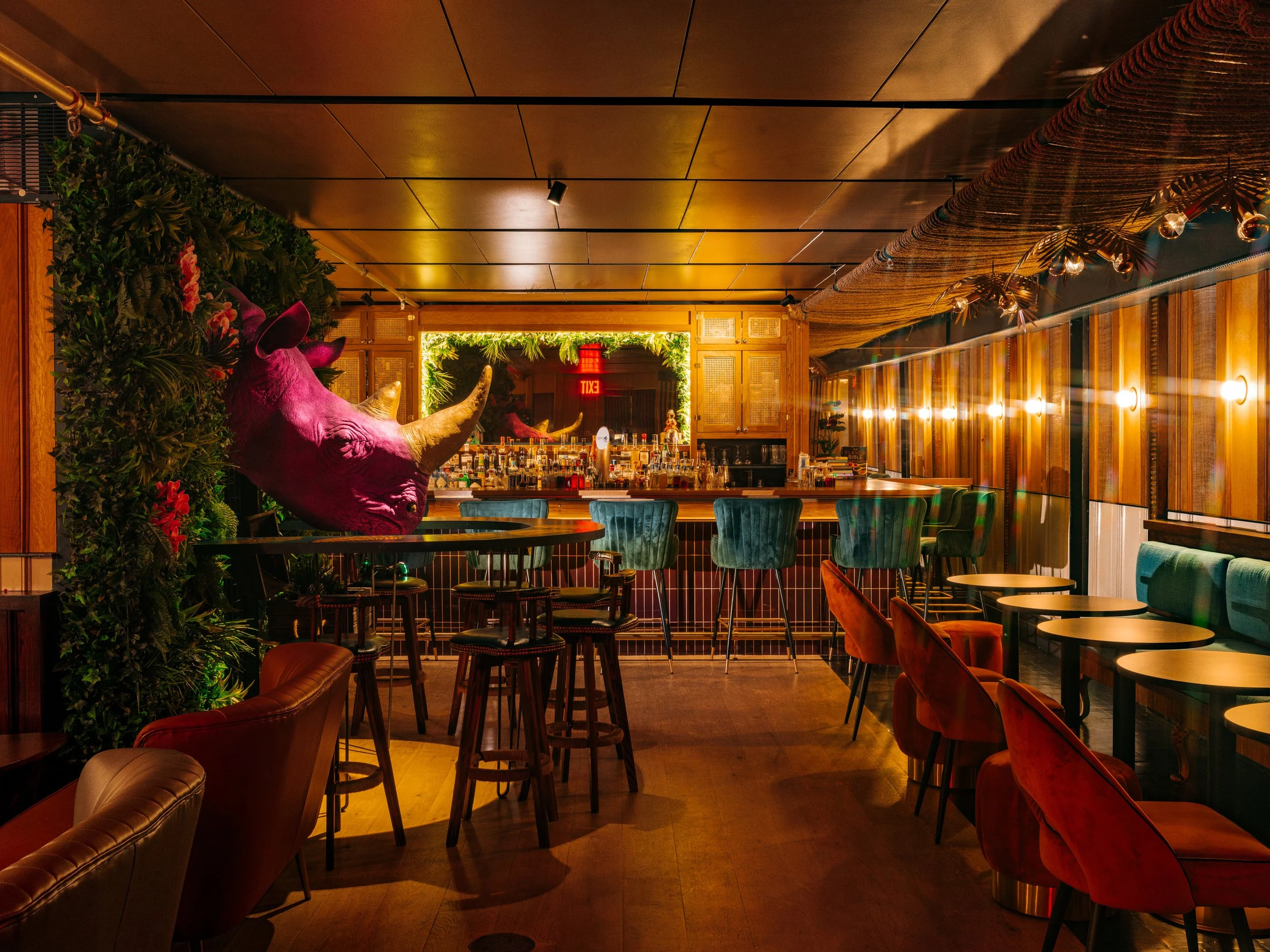  jewel-toned lit bar with giant pink rhinoceros sculpture in the middle of the room with a round bar surrounding it and green leafy accents around mirror behind bar 