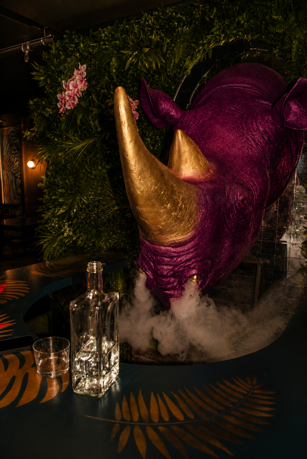  pink rhino sculpture with gold horns blowing smoke out of its nostrils towards a clear bottle of liquor 
