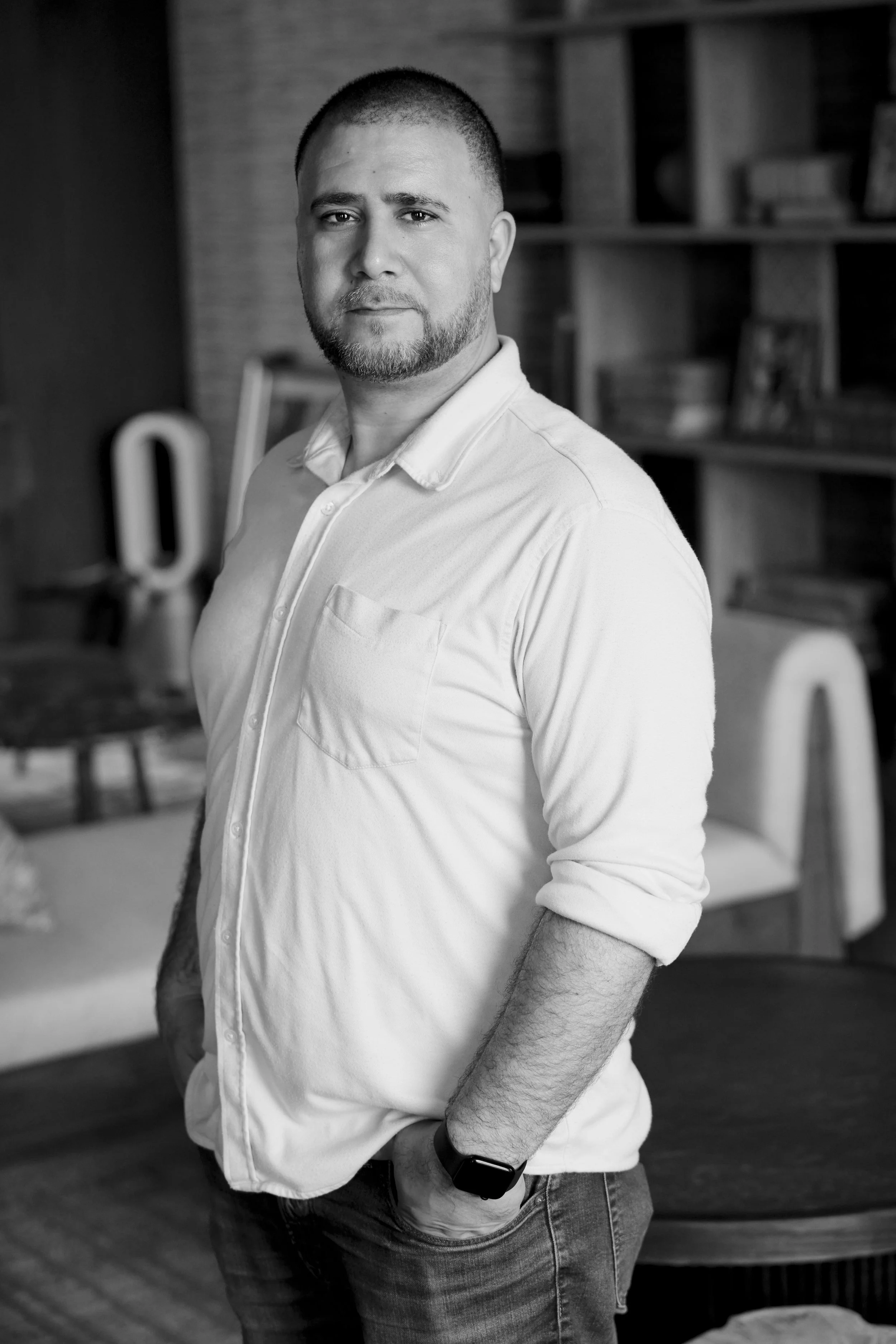 A man with a beard and short hair, wearing a white button-up shirt and jeans, standing indoors with bookshelves and a Dyson vacuum cleaner in the background.