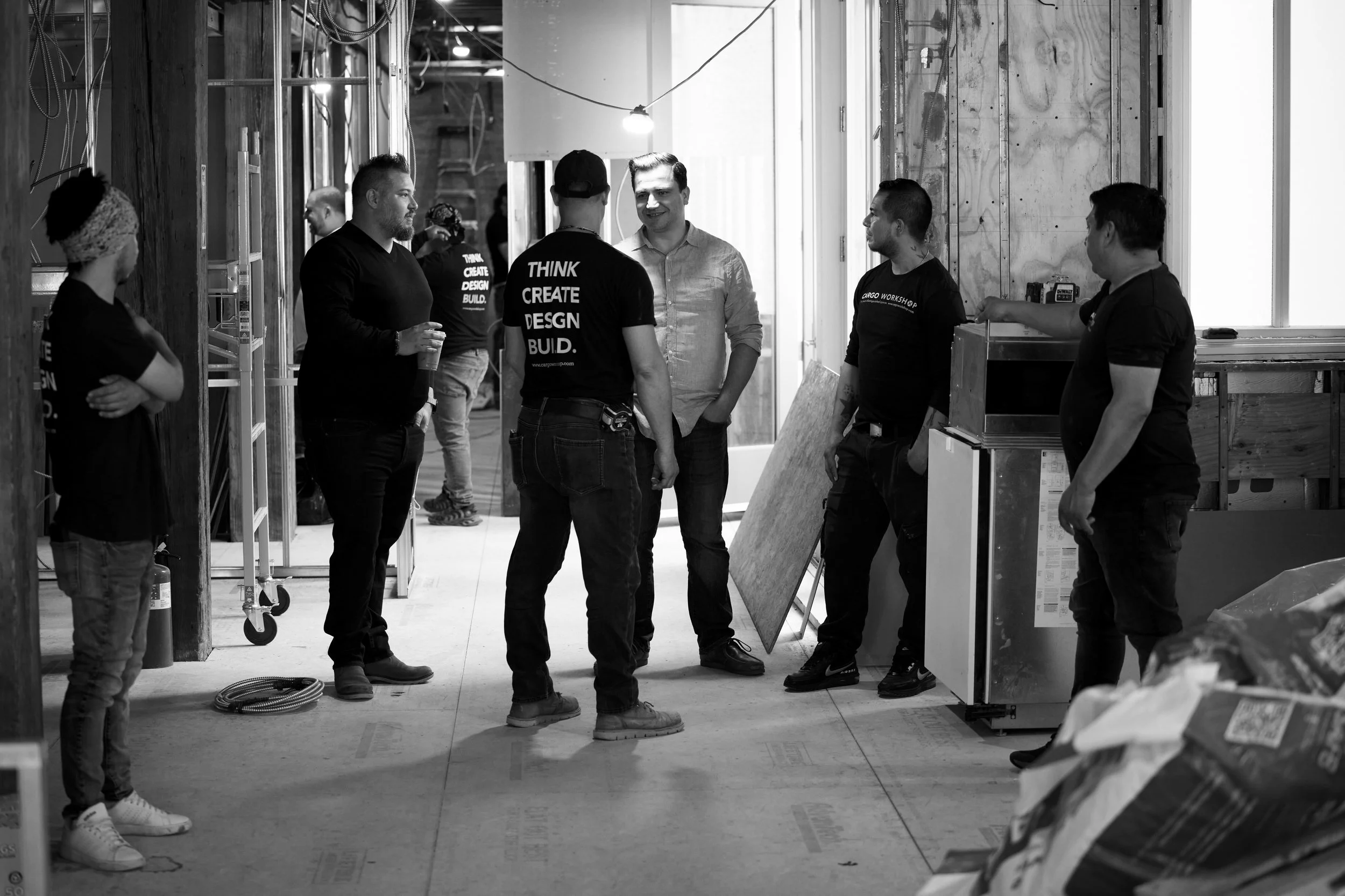 A group of people inside a construction site, some wearing black T-shirts, engaging in conversation, with exposed wooden framing and electrical wiring in the background.