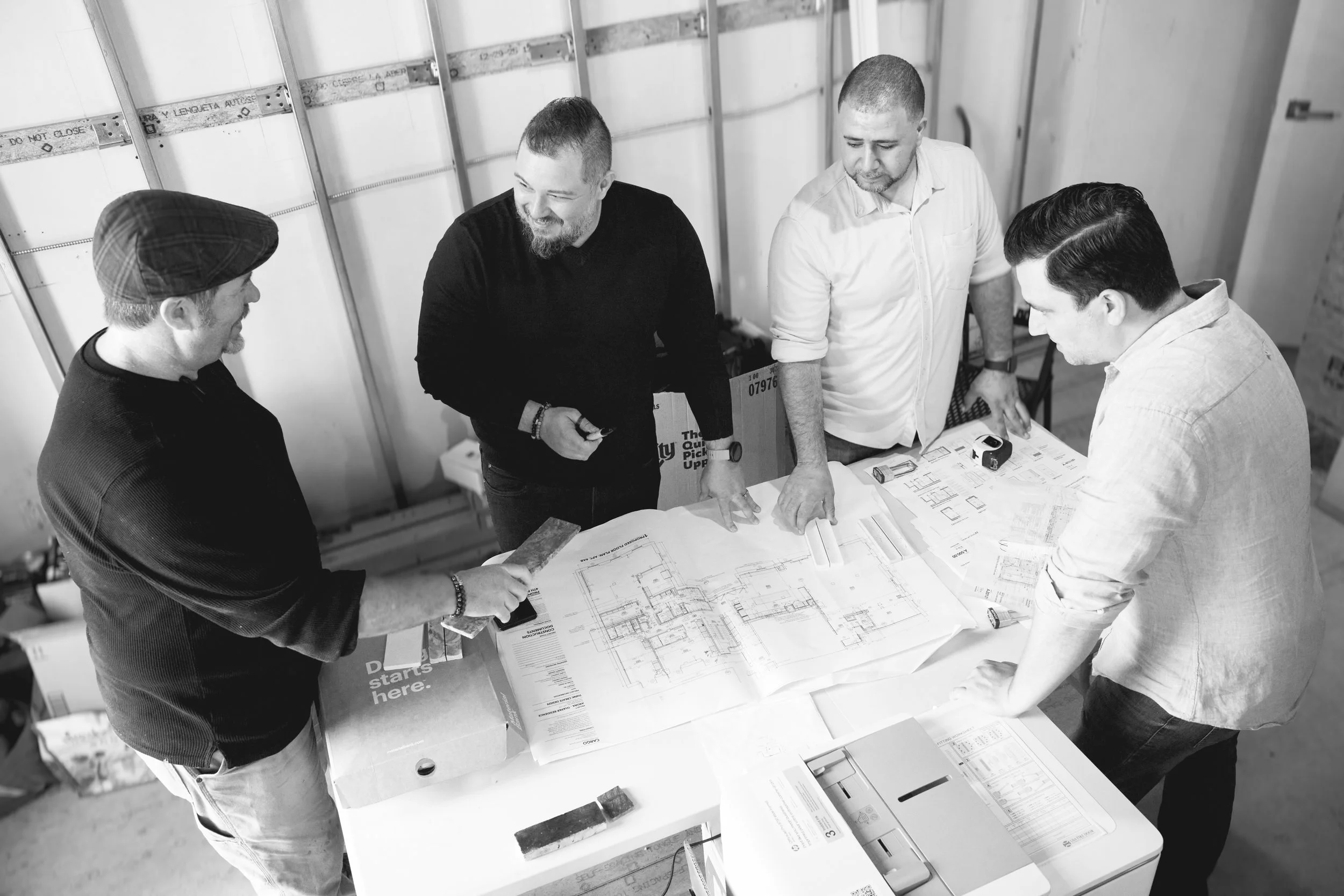 Four men are gathered around a table reviewing architectural floor plans in a construction site or warehouse. They appear to be discussing the plans, with one man pointing at the blueprints, and the others listening and looking at the plans.