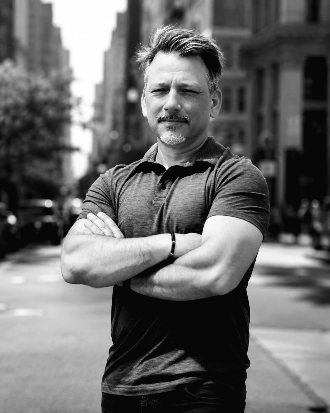 A man standing outdoors on a city street with his arms crossed. He has short hair, a goatee, and is wearing a casual polo shirt.