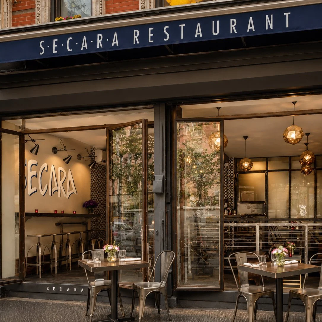 Exterior view of Seca R A restaurant with outdoor seating on a city sidewalk, featuring tables with flowers, glassware, and chairs, and a partially open entrance leading to a cozy interior.