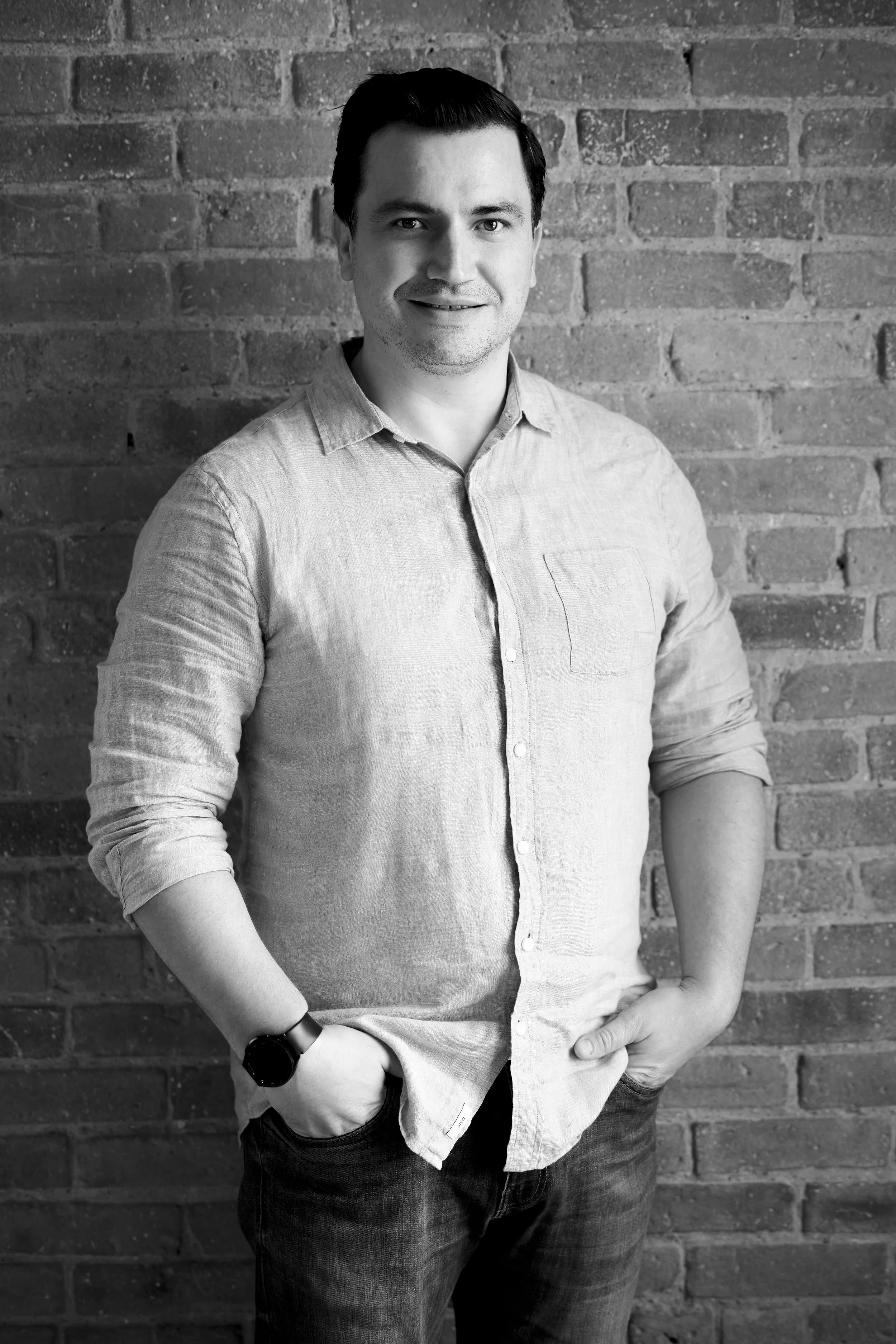 A man with dark hair, wearing a light-colored button-up shirt and dark jeans, standing against a brick wall, smiling with one hand in his pocket.