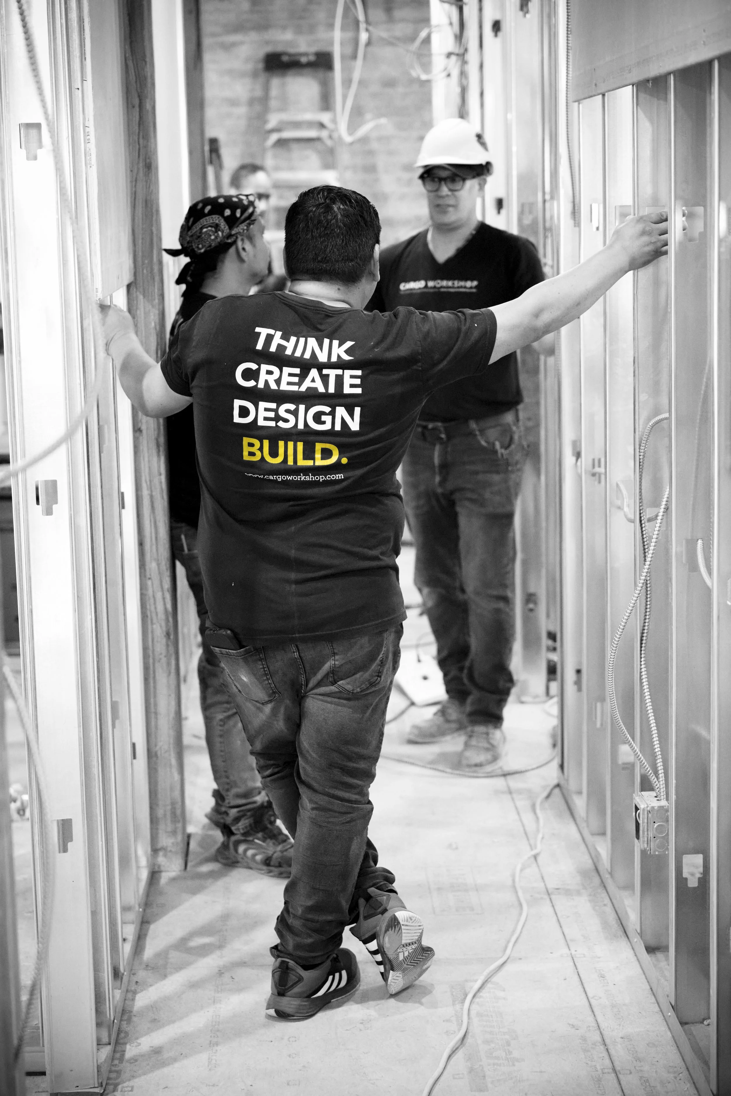 Four construction workers inside a building under construction, with exposed wall framing and electrical wiring. One worker is wearing a shirt with the words 'Think, Create, Design, Build' on the back. Everyone is wearing safety gear including helmets and glasses.