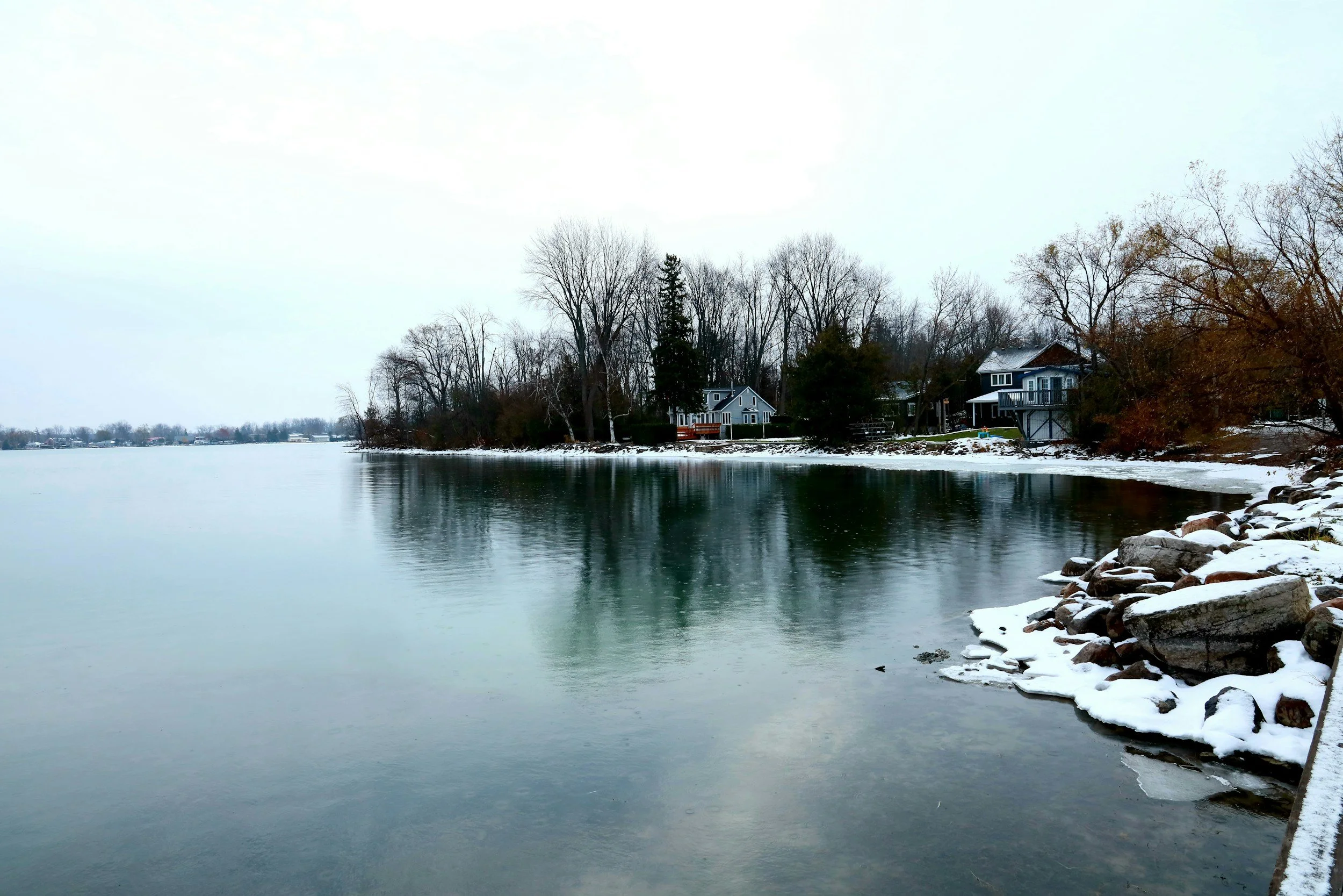 Snow-covered shoreline with rocks and trees, houses in the background, and a calm body of water reflecting the landscape
