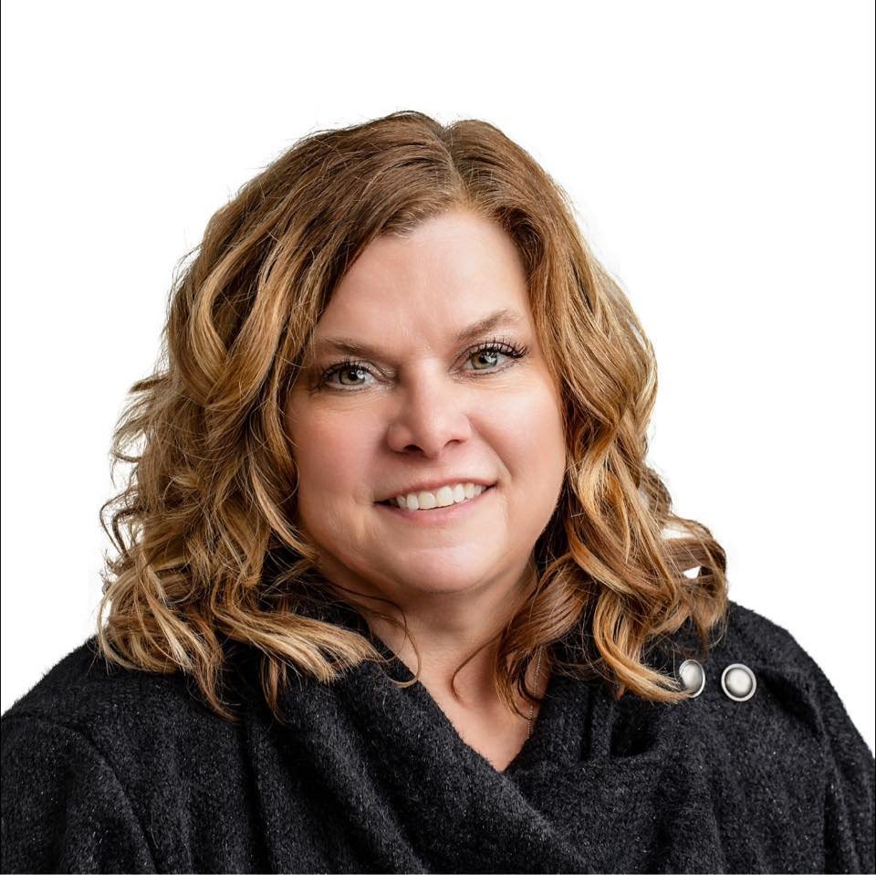 A woman with shoulder-length wavy red hair smiling against a white background, wearing a black top with decorative buttons.
