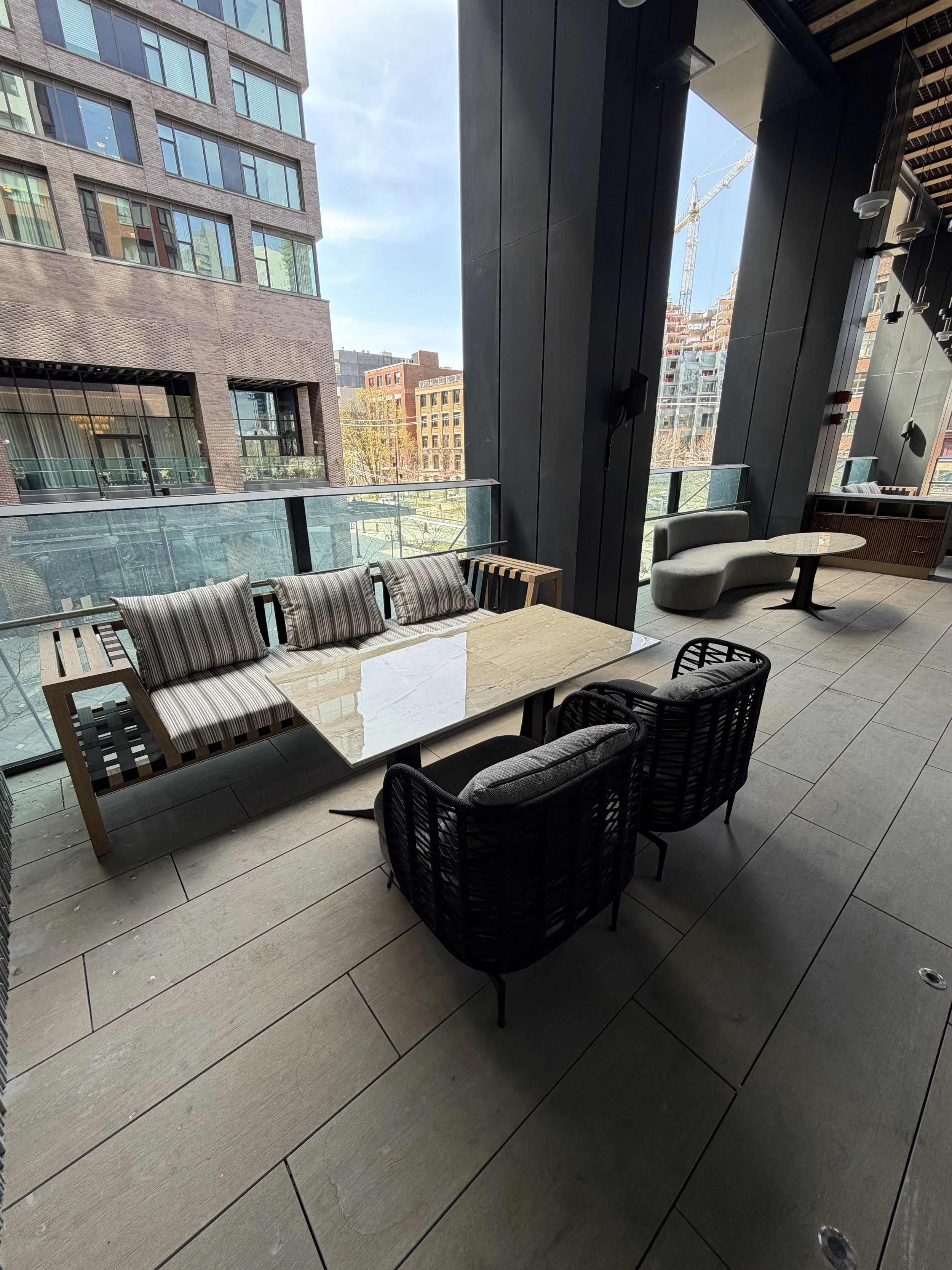 Indoor seating area with a marble table, black woven chairs, and a curved gray ottoman with a small round table. Large windows overlook a cityscape with tall buildings and construction cranes. The indoor area has dark walls and tiled flooring.