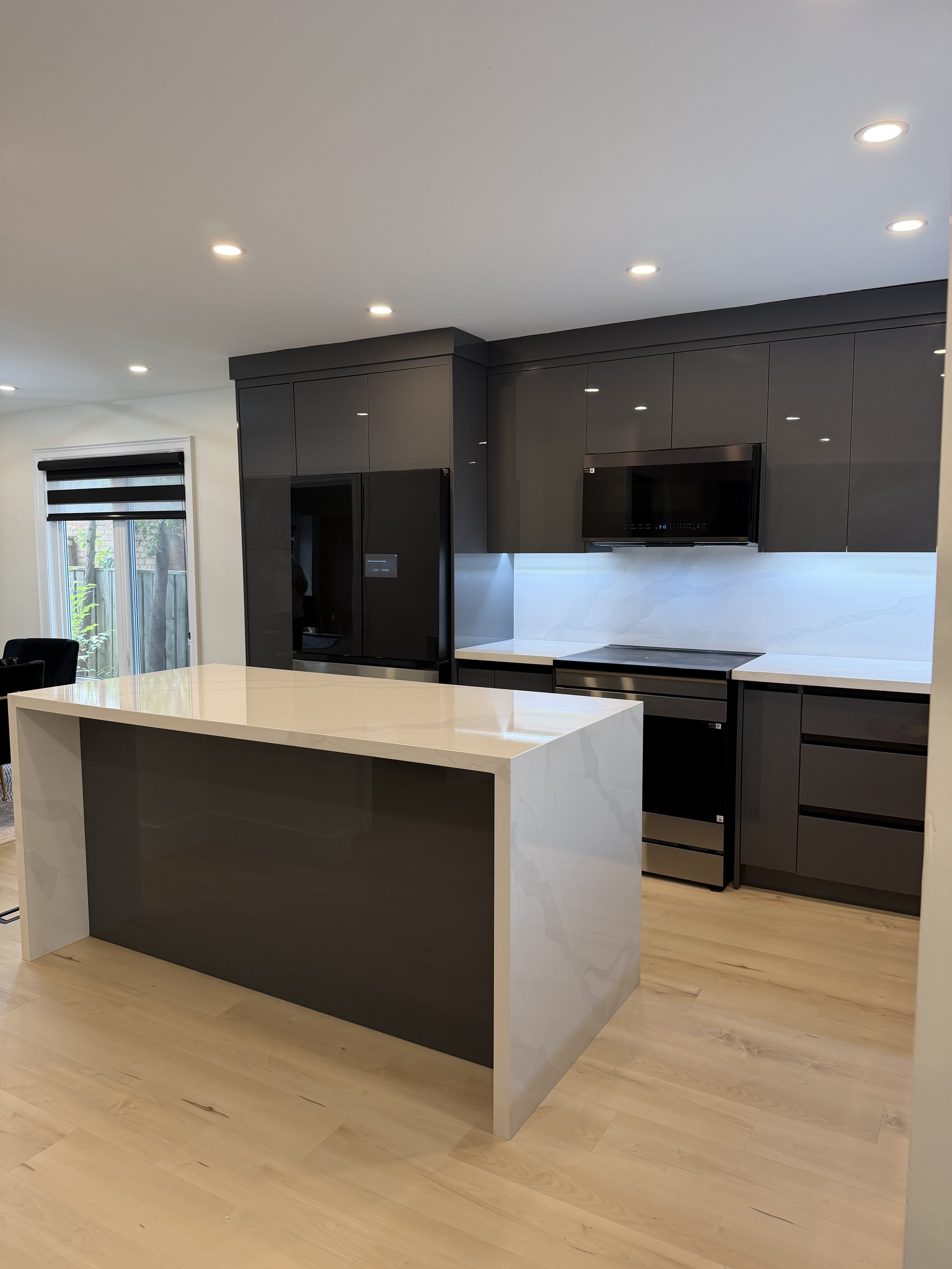 Modern kitchen with black cabinetry, built-in appliances, a white marble island, light wood flooring, and a sliding glass door with blinds.