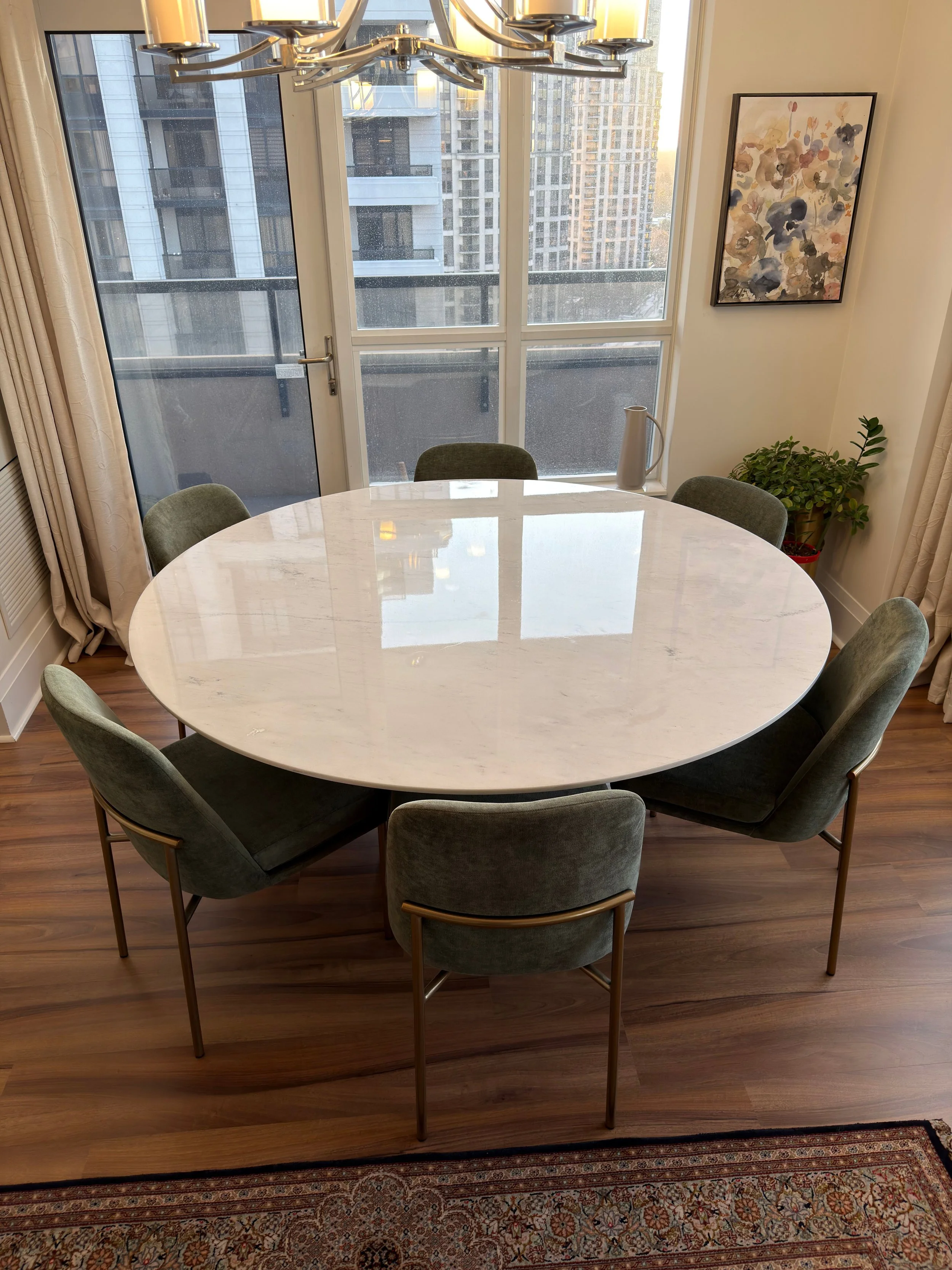 A modern dining room with a round white marble table, six green upholstered chairs, a chandelier overhead, a large glass door leading to a balcony, a plant in a pot, a colorful abstract painting, and a patterned rug on a wooden floor.