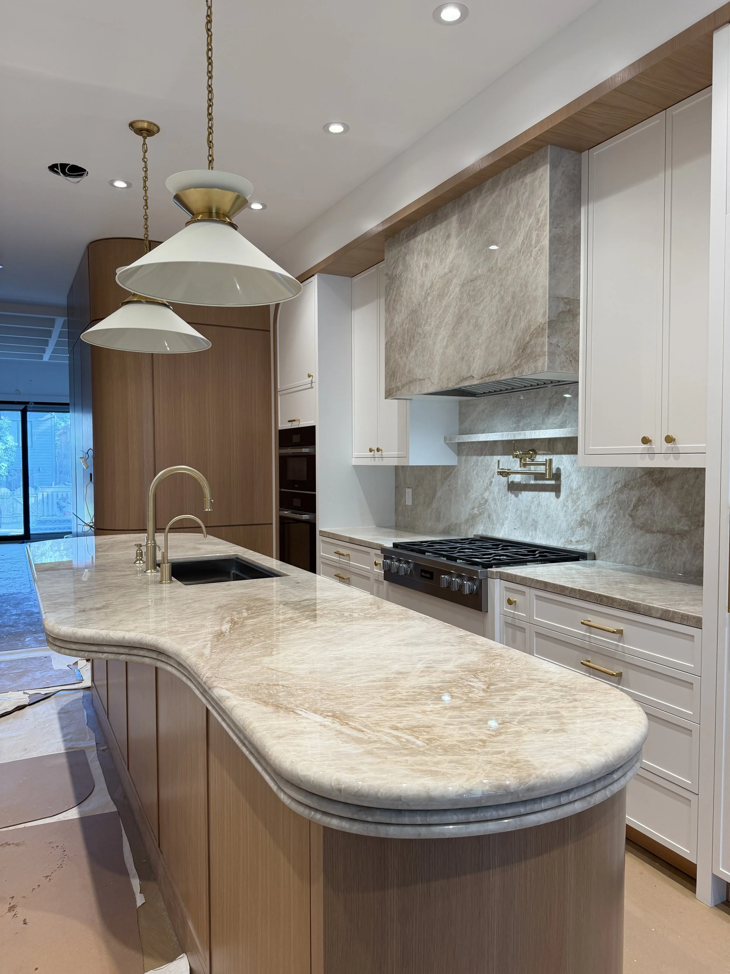 Modern kitchen with a rounded beige marble countertop island, white cabinets with gold handles, a beige marble backsplash, and a gold-brushed kitchen faucet. Pendant lights hang from the ceiling.