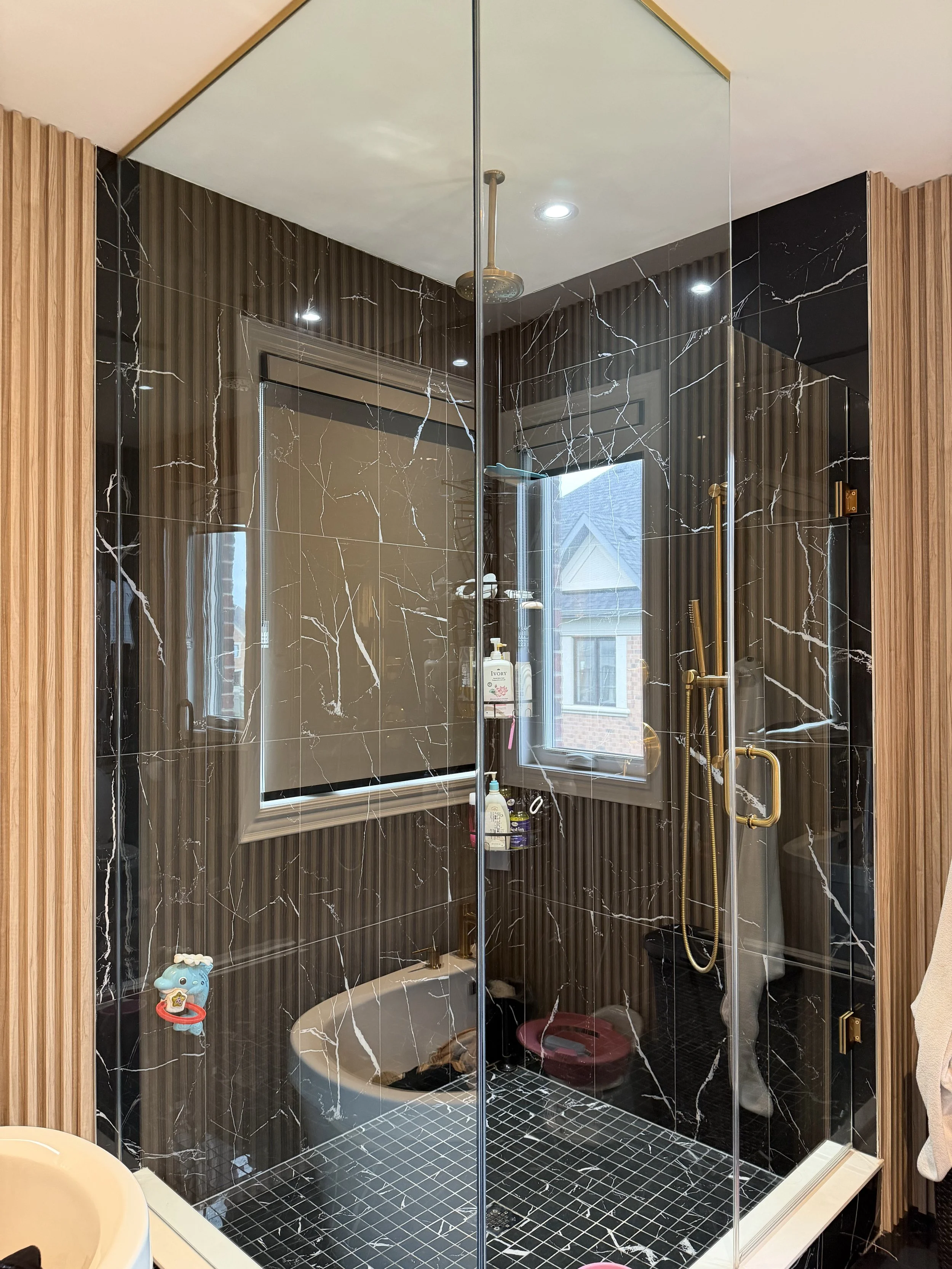 Modern walk-in shower with black marble walls and gold fixtures, including a rain showerhead, a handheld shower, and a window with outside view.