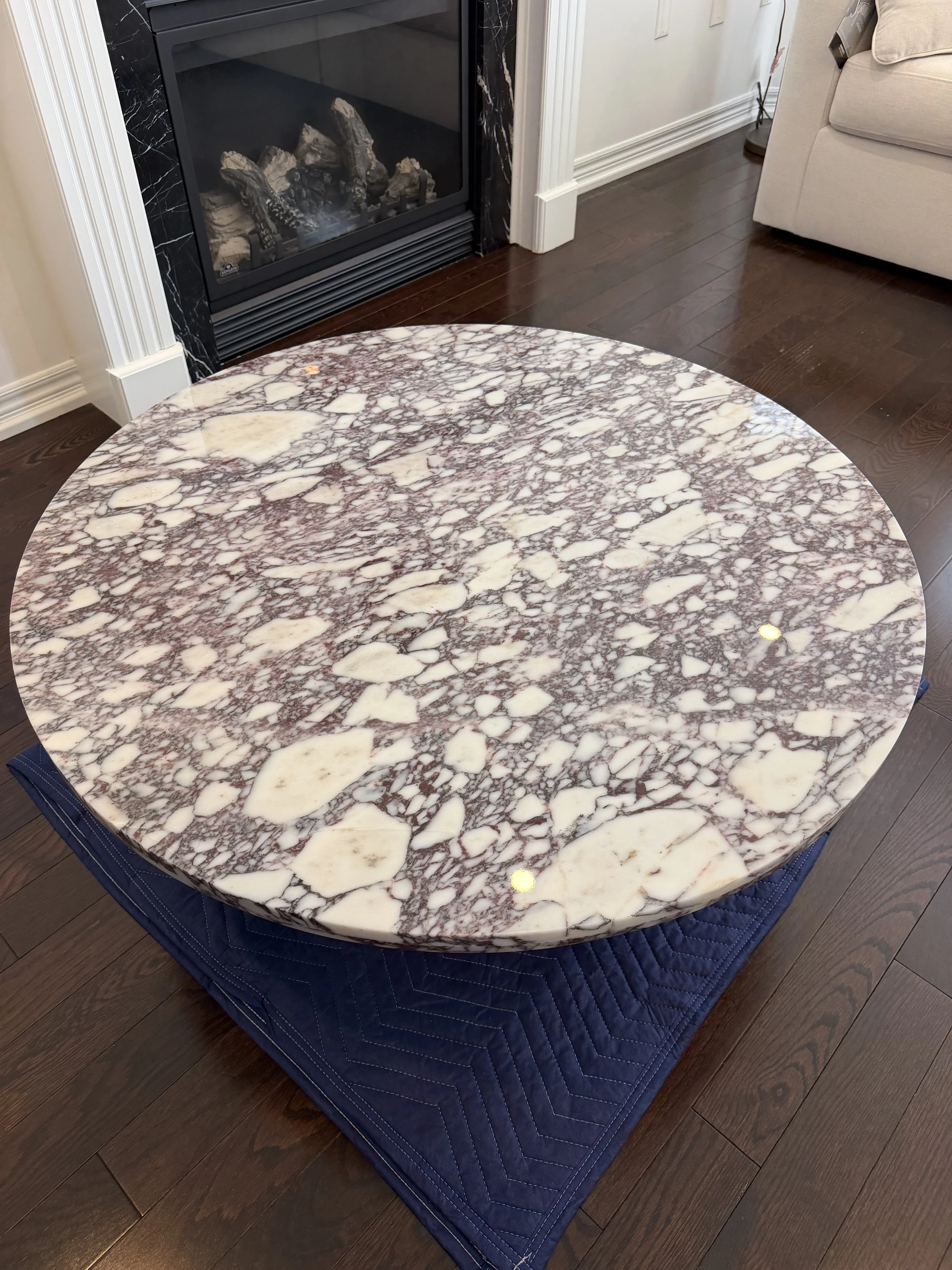 Close-up of a round marble table with white and brown veining, positioned on a blue quilted tablecloth on a dark hardwood floor, near a fireplace and beige sofa.