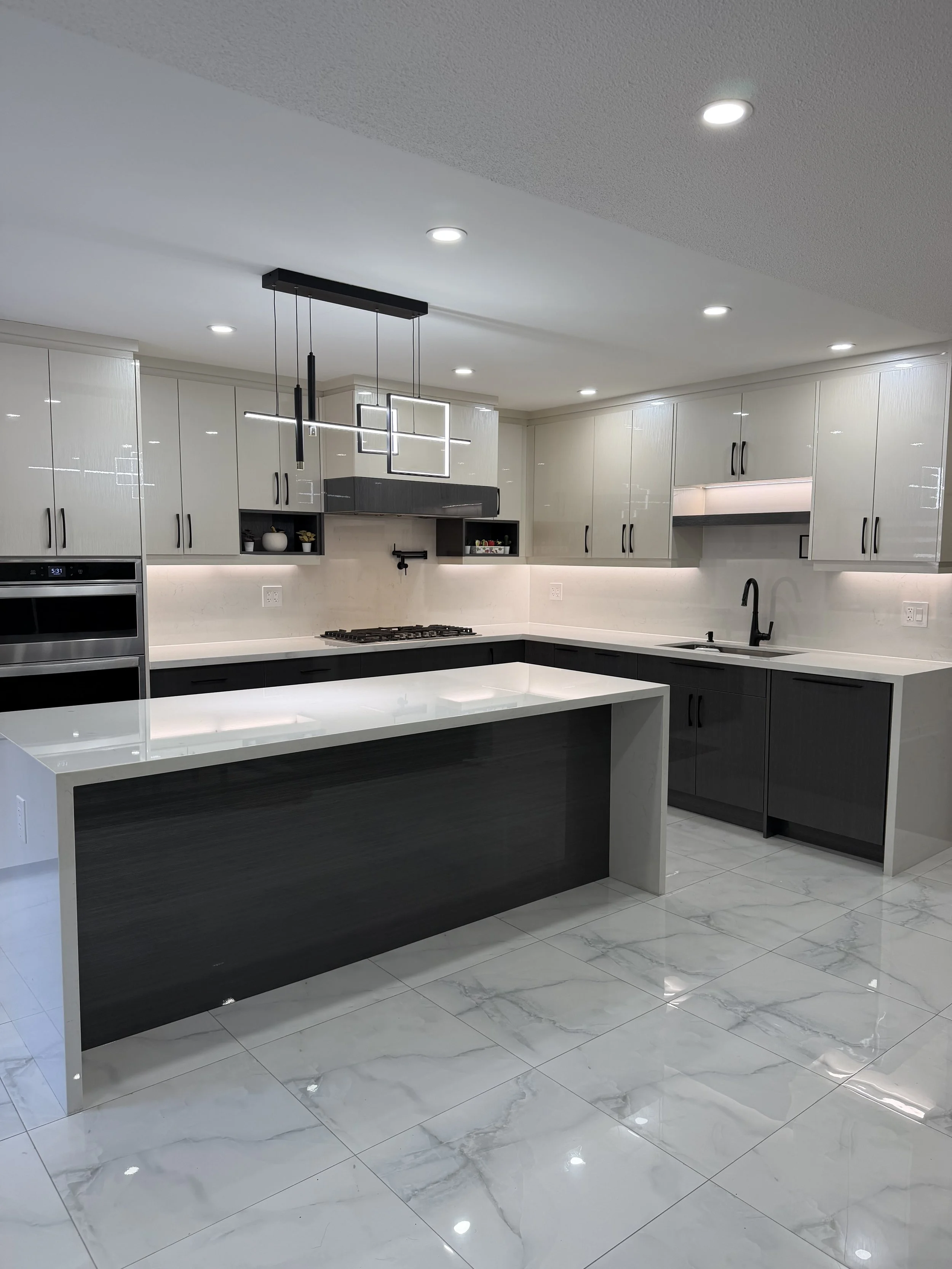 Modern kitchen with white and dark gray cabinets, marble tile floor, and a black and white island countertop. Recessed ceiling lights and a contemporary light fixture hang over the island. Sink with black faucet is visible in the corner.