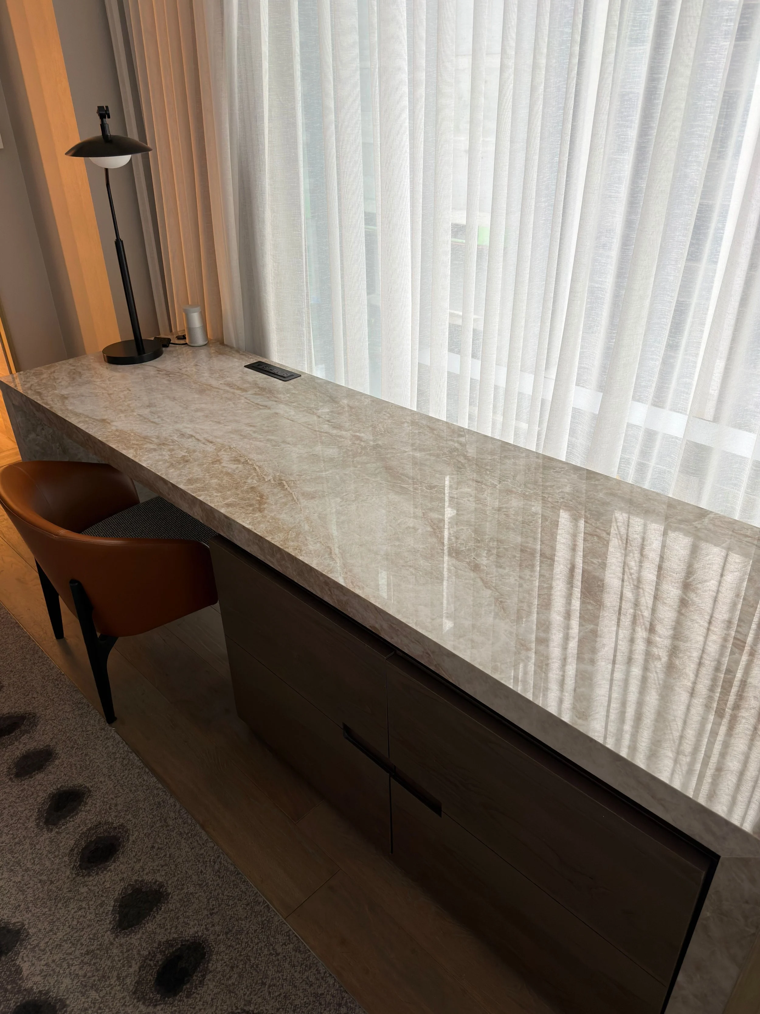 A marble countertop with a black desk lamp, a small speaker, a remote control, and a brown chair in front of a large window with sheer white curtains.