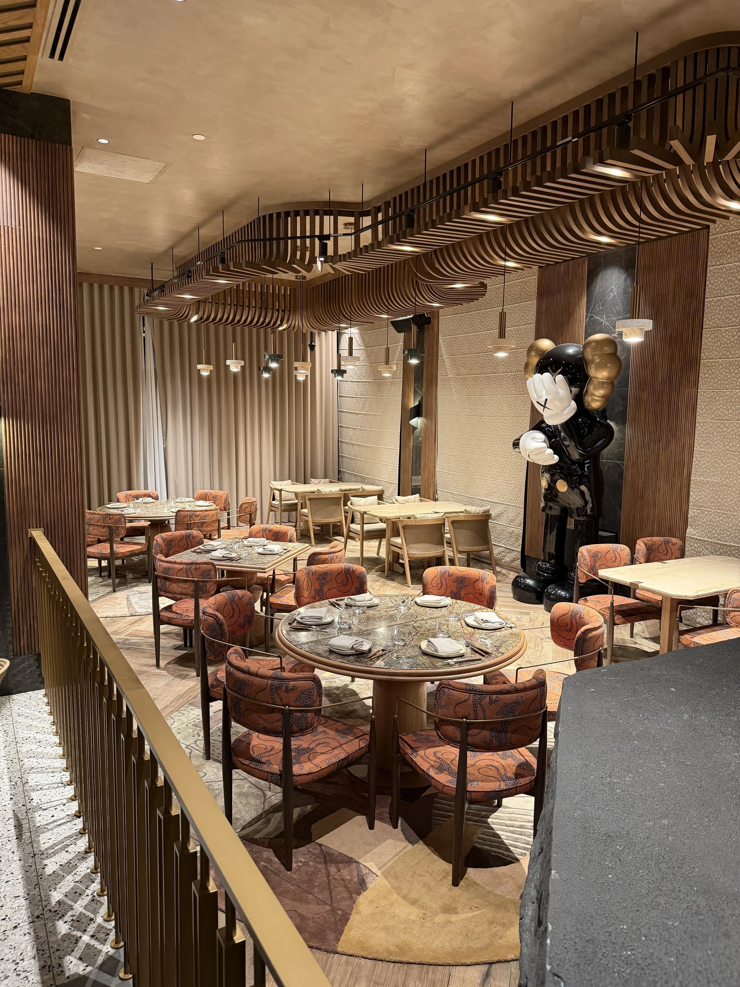 Interior of a restaurant or hotel dining area with tables, chairs, and decorative lighting, featuring a modern design with wood accents and a large artist sculpture of a cartoon character.