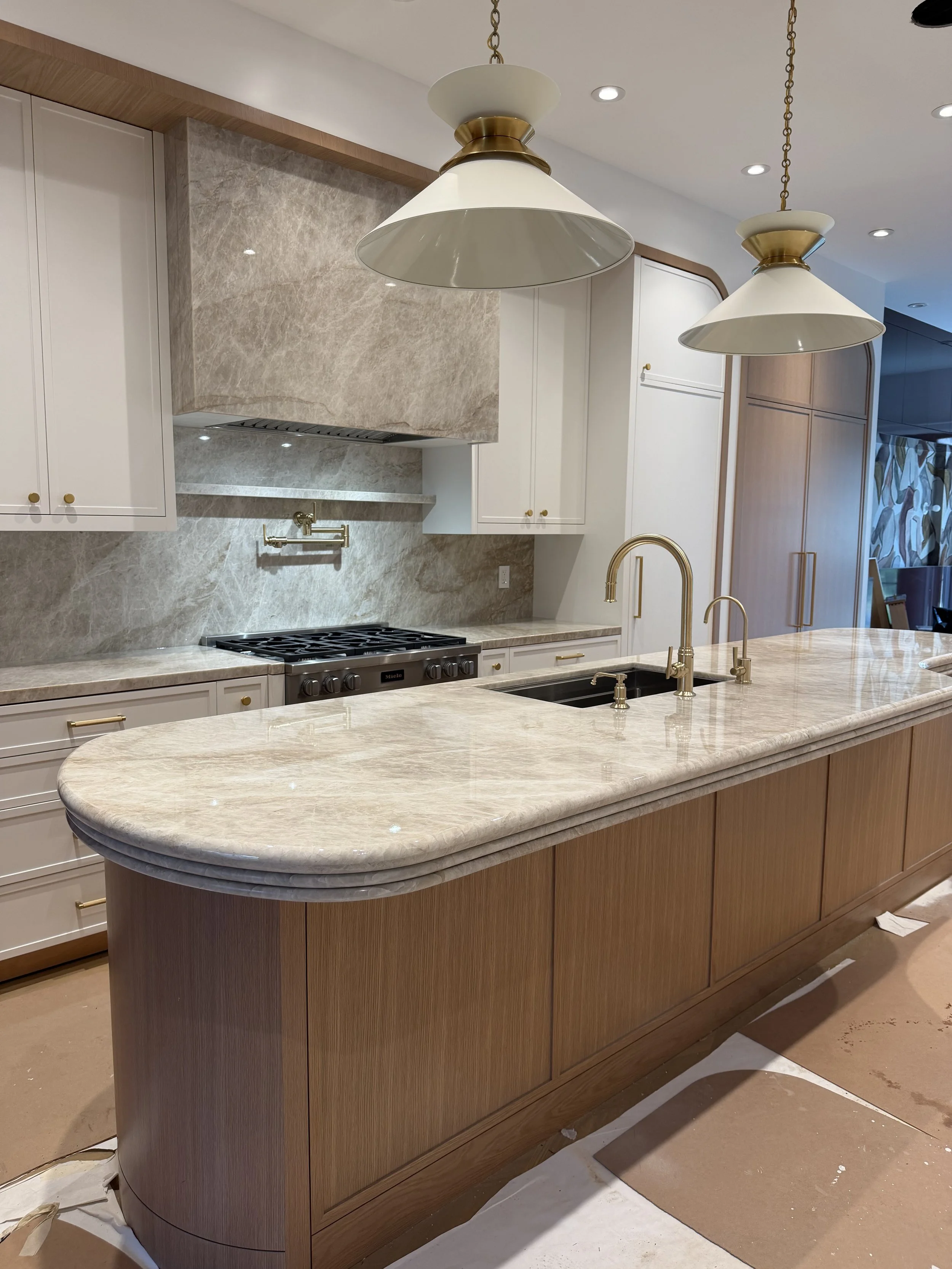 A kitchen island with a marble countertop, built-in sink, and gold fixtures. White cabinets with gold knobs, a marble backsplash, and pendant lights with gold accents hanging from the ceiling.