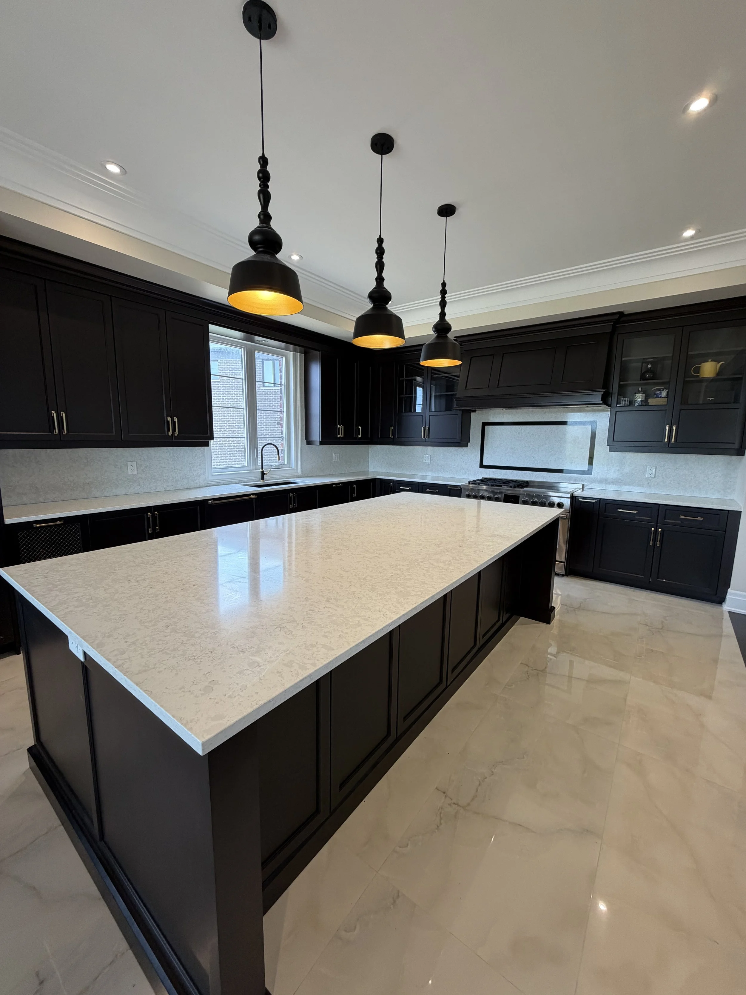 Modern kitchen with black cabinets, white granite island, tiled floor, and three black pendant lights hanging from the ceiling.