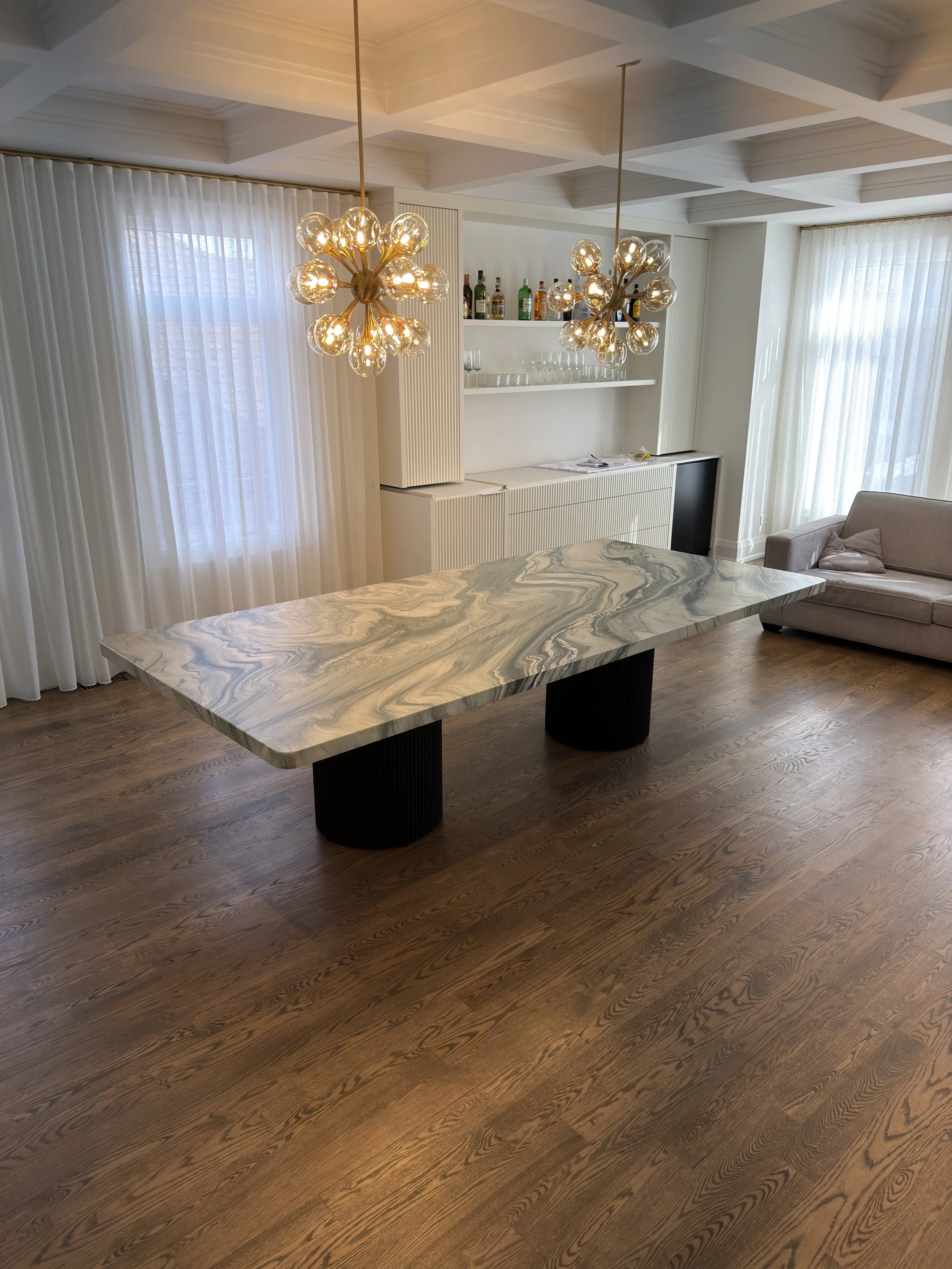 Modern living room with a large marble table, two gold chandelier lights, white curtains, and a bar shelf with bottles and glasses.