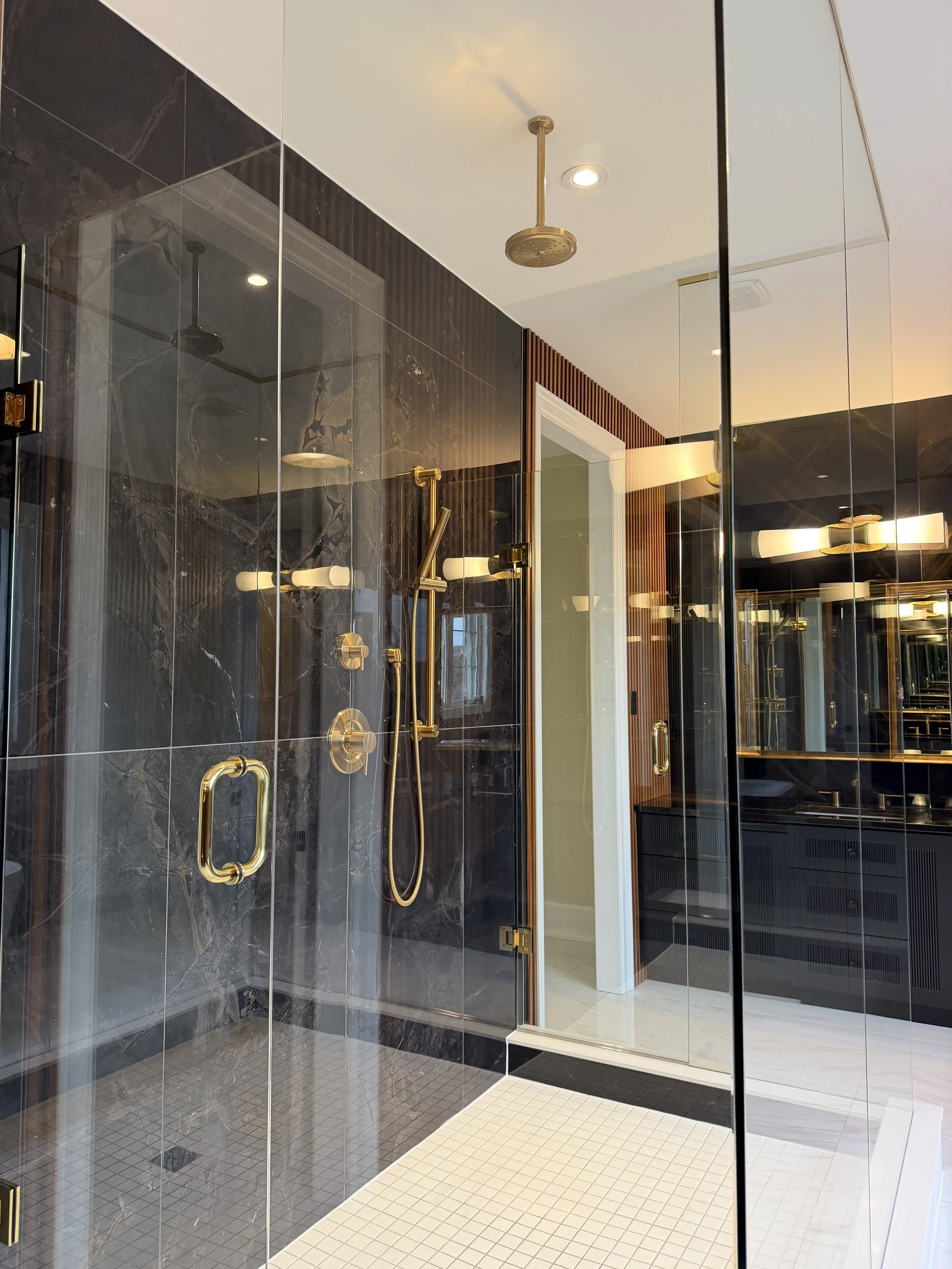 A modern bathroom with a walk-in shower enclosed by glass panels, featuring black marble walls, gold fixtures, a rainfall showerhead, and a separate handheld shower, with a white tiled floor and a wooden slatted wall and a black cabinet in the background.
