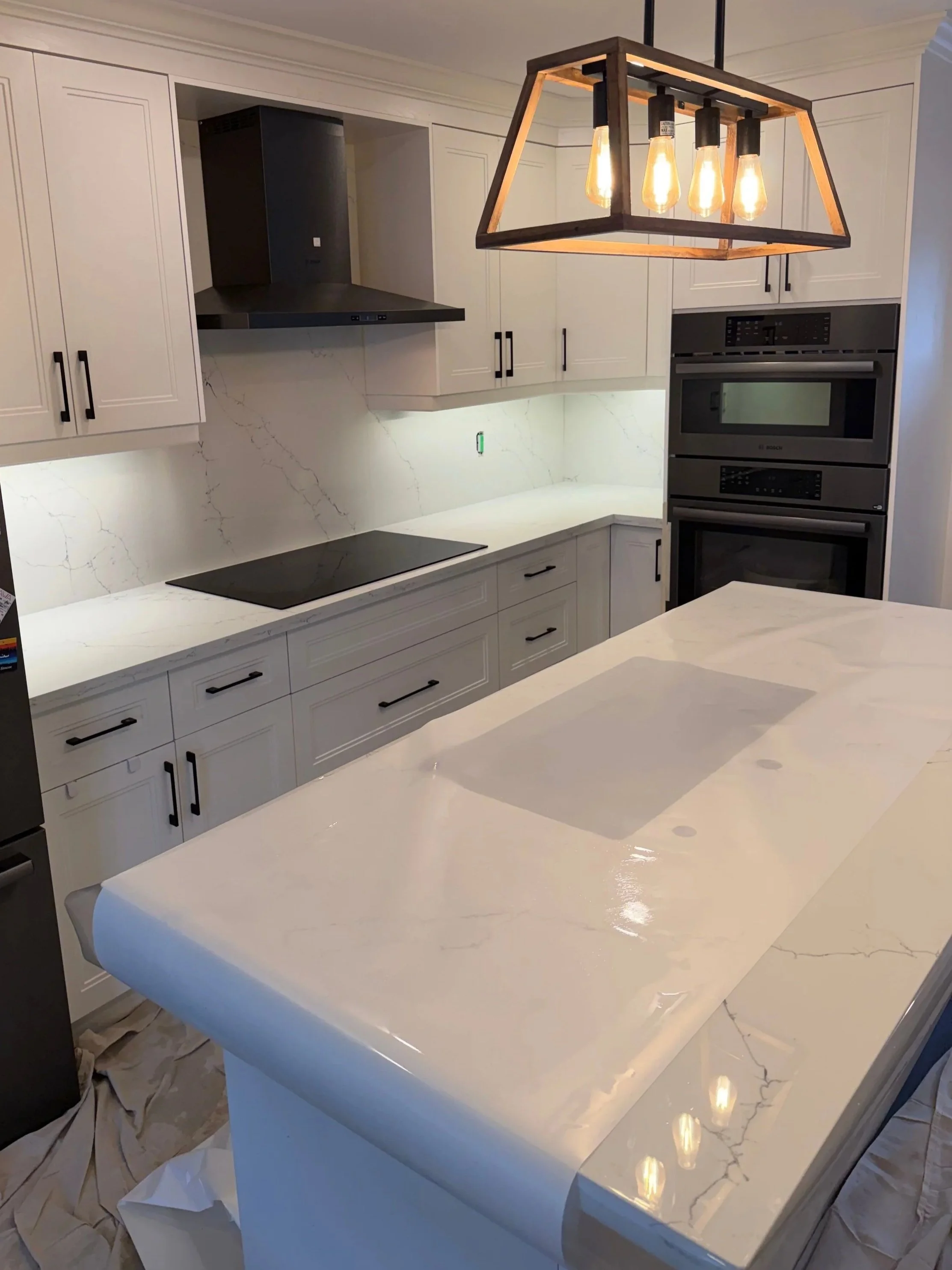 Modern kitchen with white cabinets, marble countertops and backsplash, black handles, black stovetop, and built-in ovens. A rectangular black and wood chandelier hangs above a white marble island.