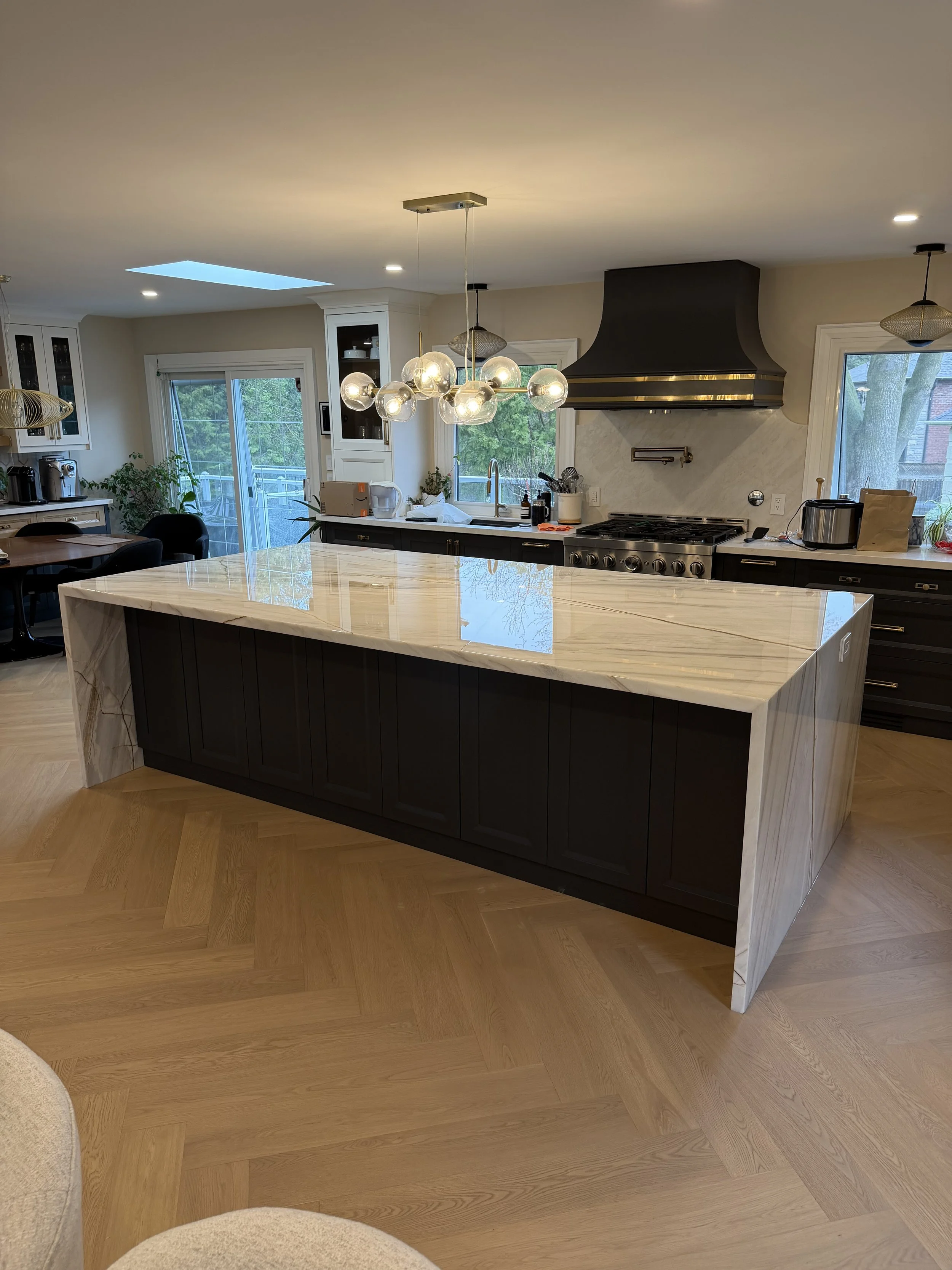 A modern kitchen with a large marble island, dark cabinets, a stainless steel range hood, pendant lights, a sliding glass door, and hardwood flooring.