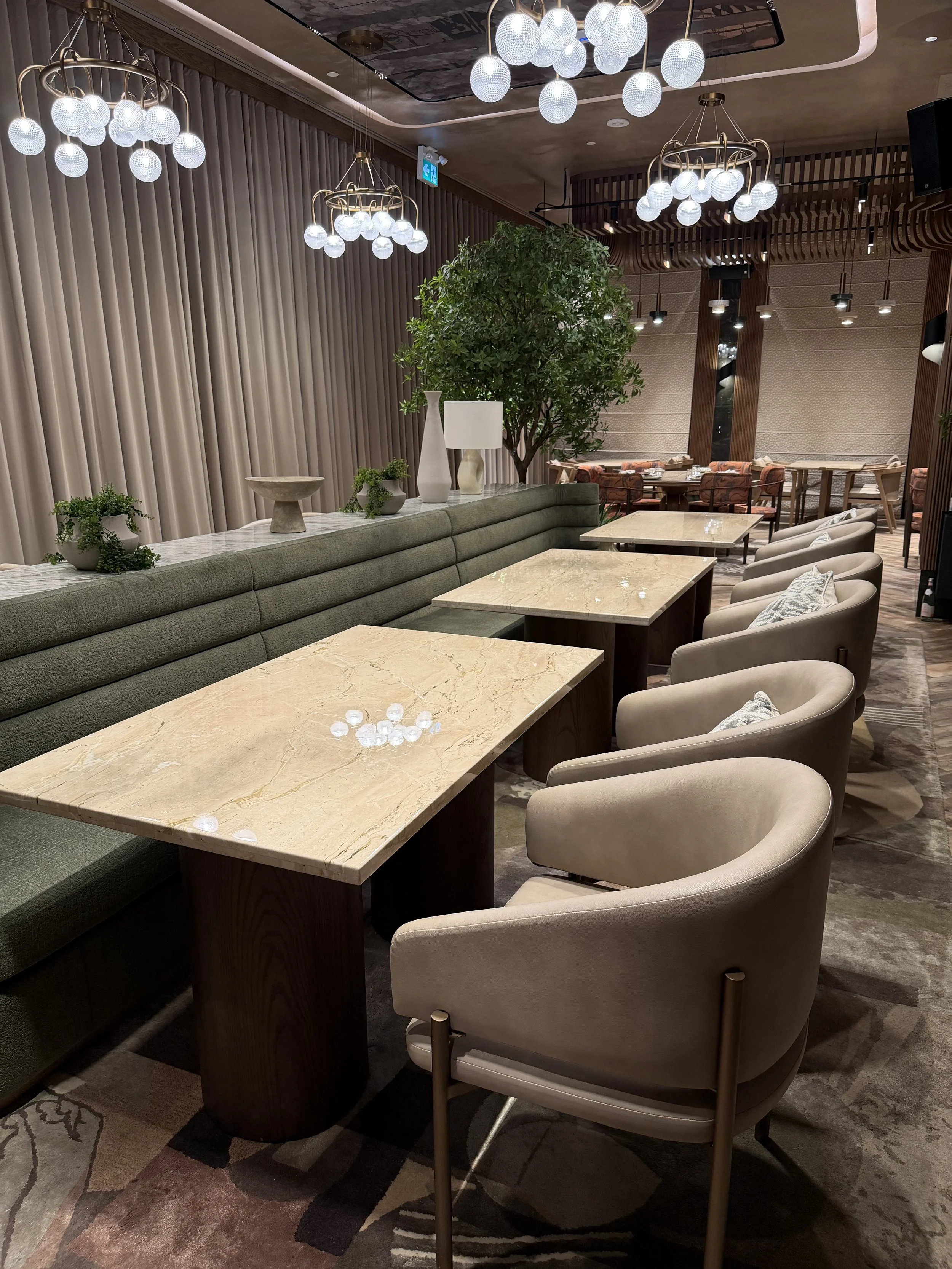 Interior of a restaurant or lounge with beige chairs, a green banquette, marble-top tables, decorative plants, large tree, and modern lighting fixtures, including globe chandeliers.