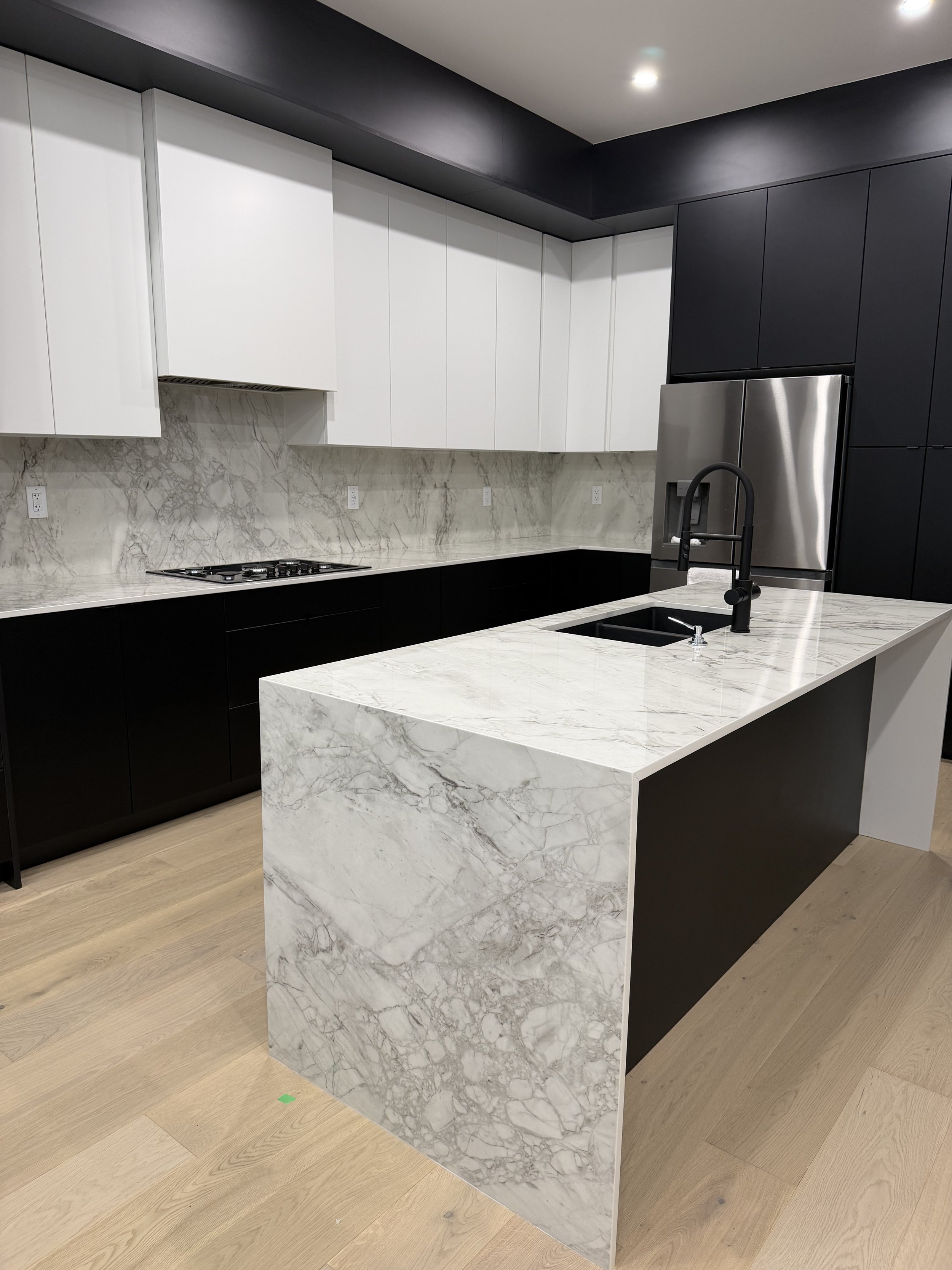 Modern kitchen with black and white cabinets, a marble island with sink, and stainless steel refrigerator, finished with light wood flooring.