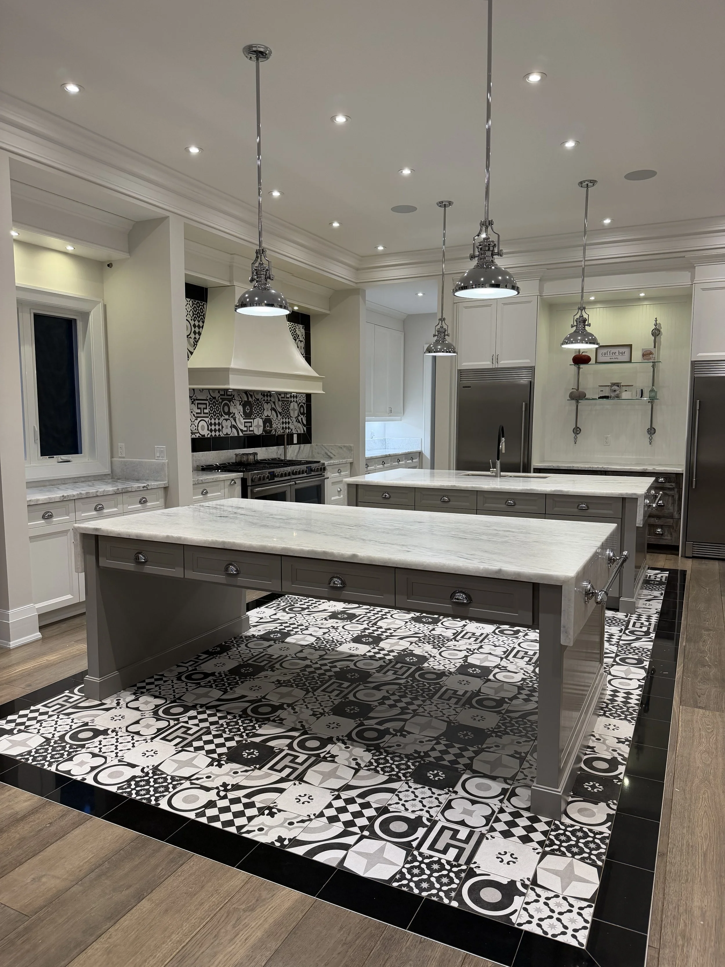 A modern kitchen with a patterned black, white, and gray tile floor, gray kitchen island, white cabinets, stainless steel appliances, and decorative shelving.