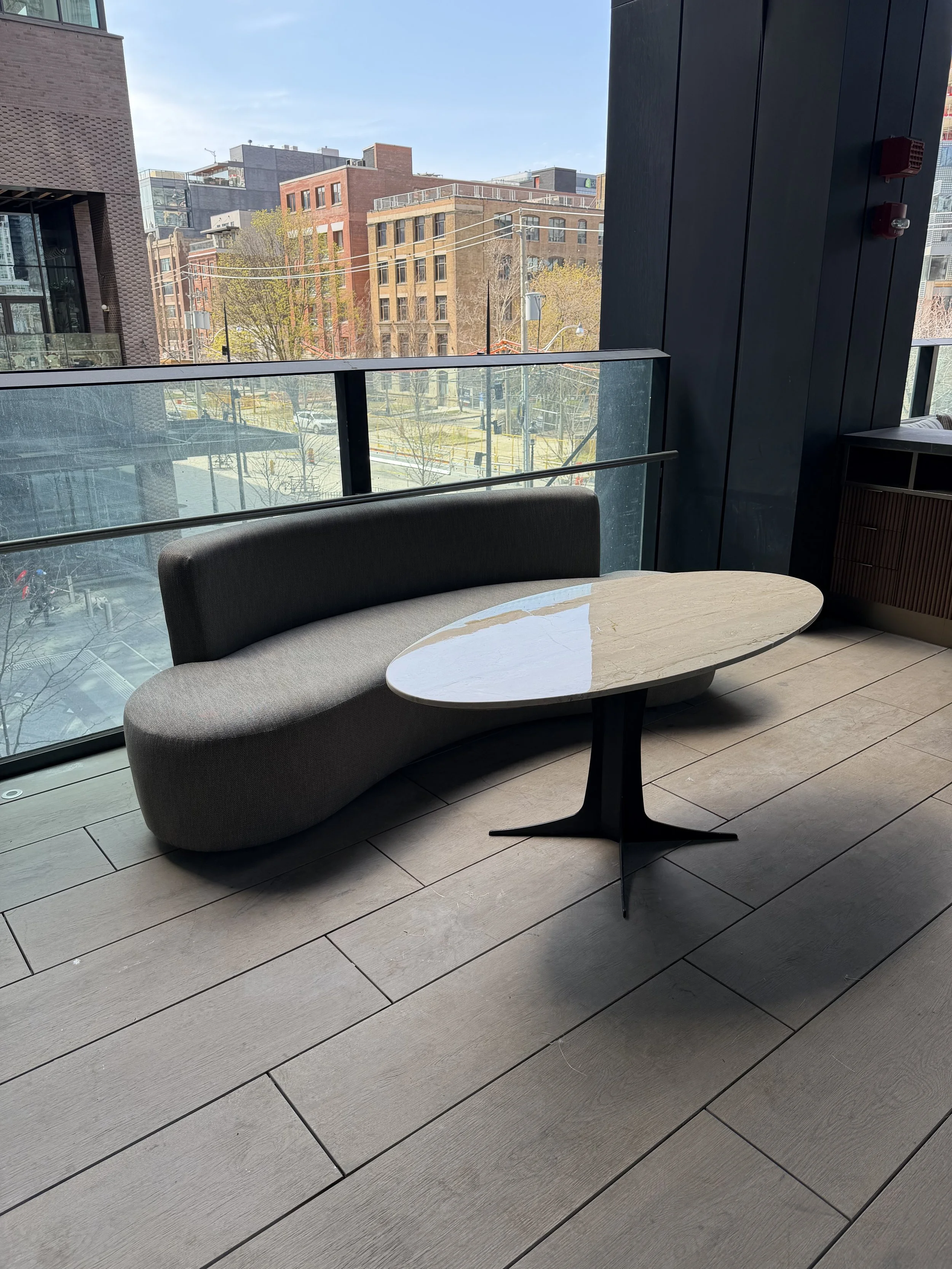 View from inside a modern building showing a seating area with a curved bench and a marble-topped table in front of floor-to-ceiling windows. Outside, city buildings, trees, and a clear sky are visible.