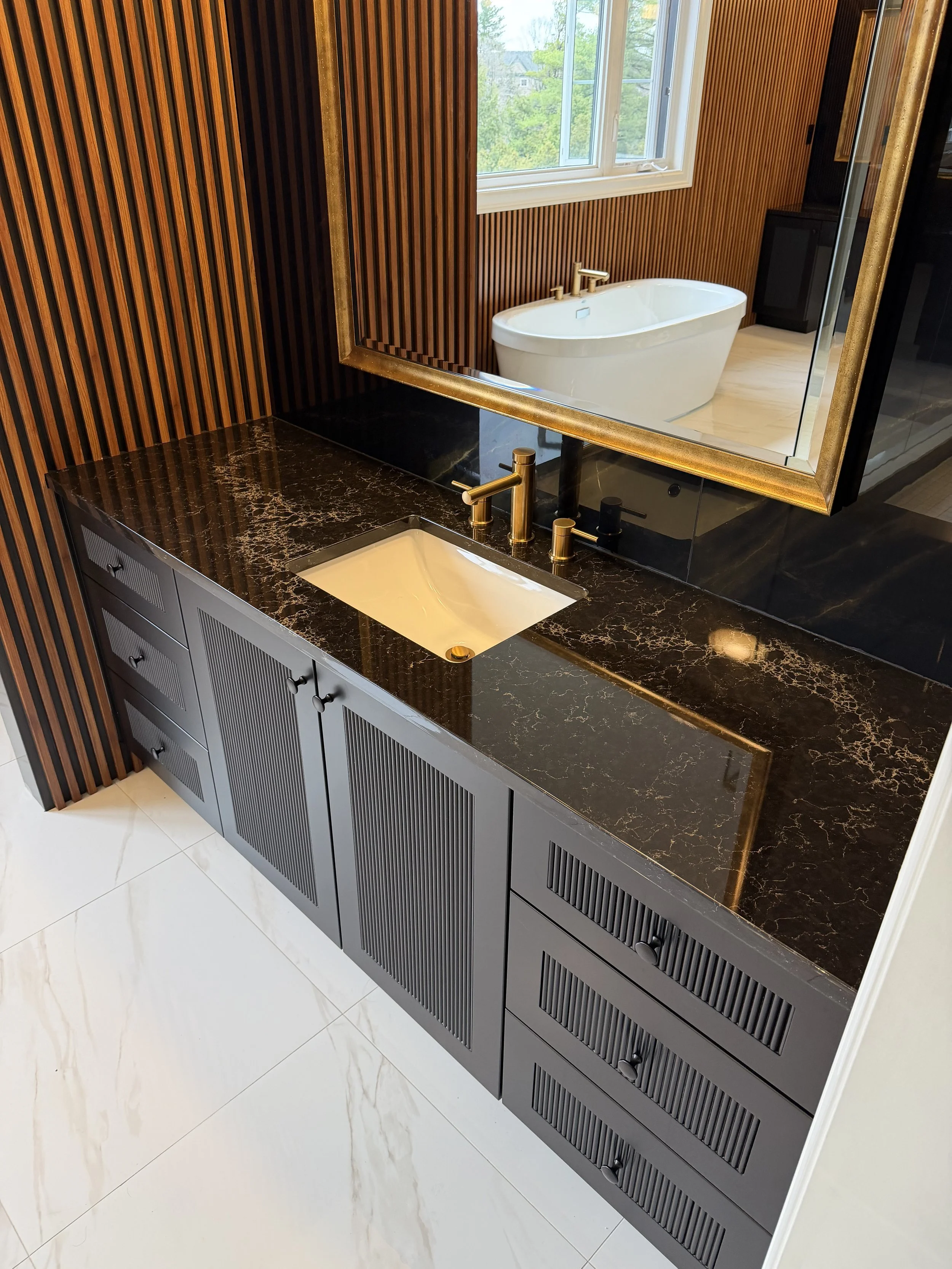 Modern bathroom vanity with a dark marble countertop, a white undermount sink, gold faucet, and black cabinet doors. Large mirror with a gold frame reflects a freestanding bathtub and a window with greenery outside. White marble floor tiles.