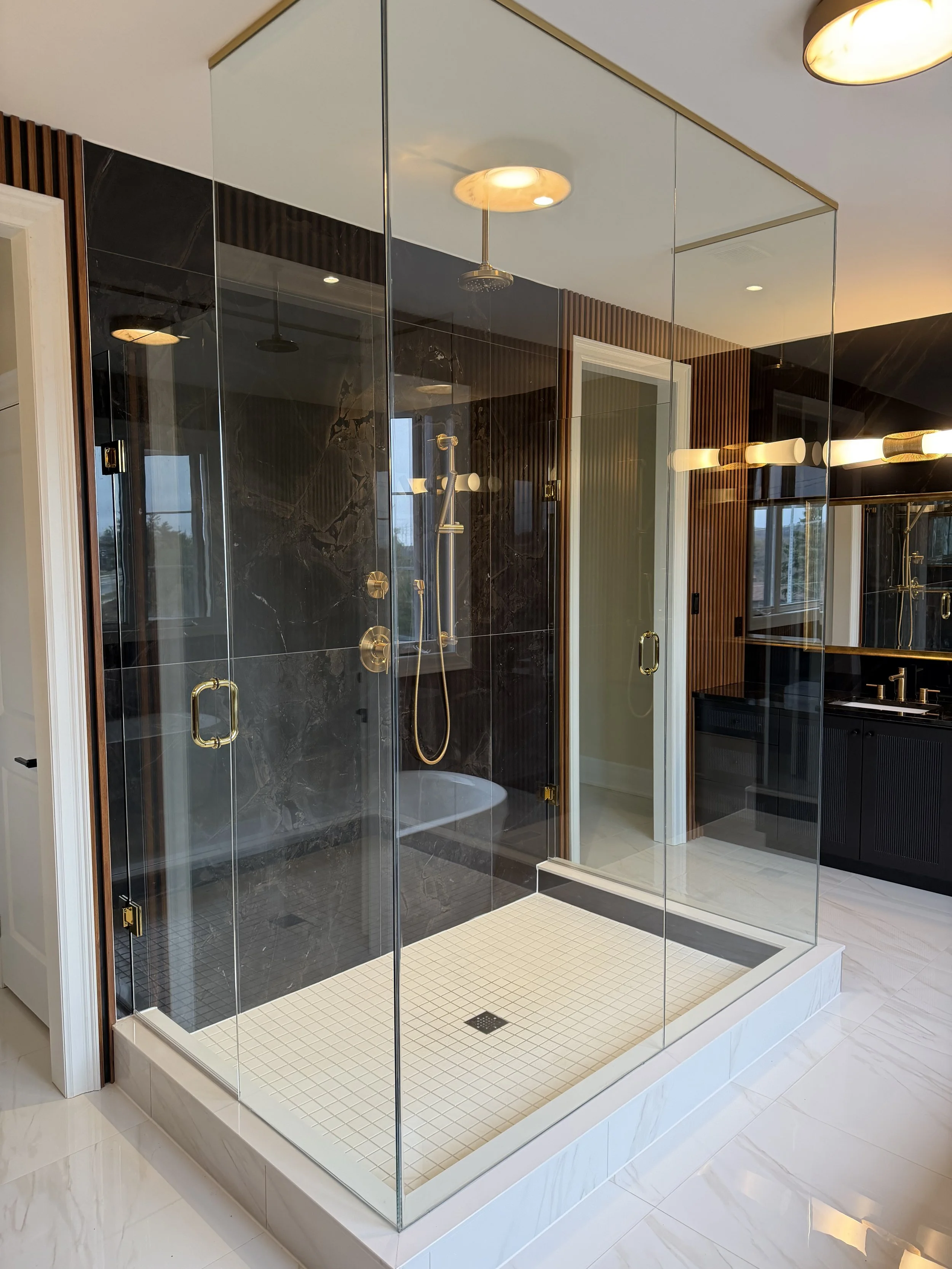 A modern glass shower enclosure with black marble walls, gold fixtures, and a built-in shower head, situated in a bathroom with white and wood accents.