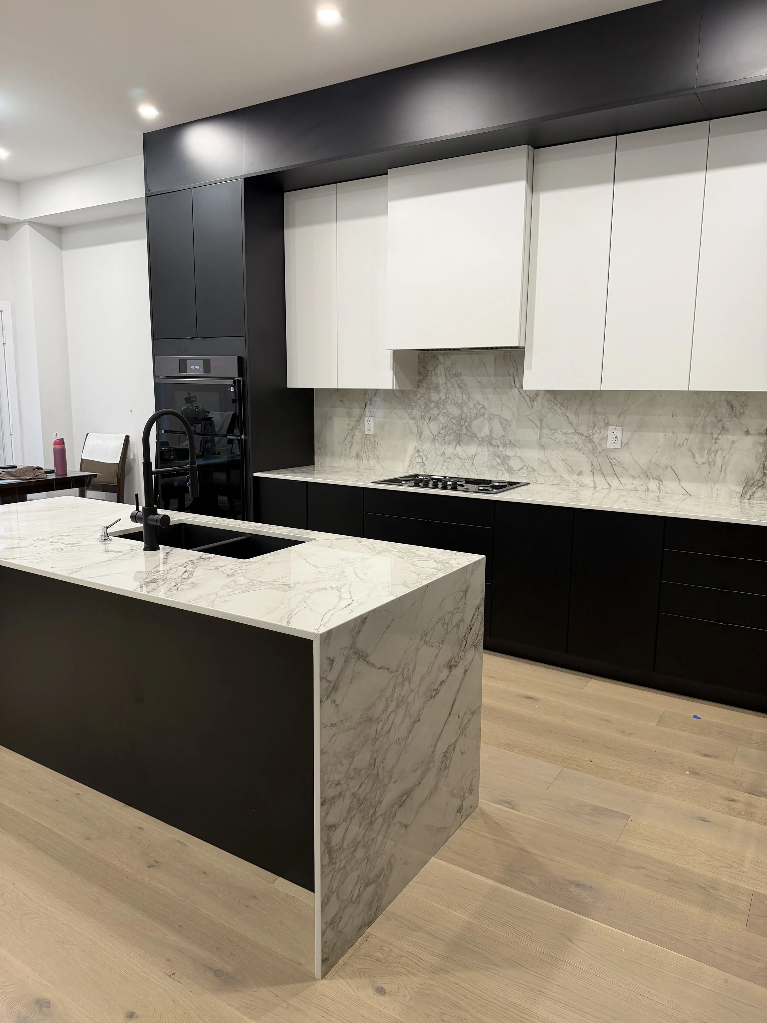 Modern kitchen with a marble island, black and white cabinets, and a stovetop, with wooden flooring and recessed ceiling lights.