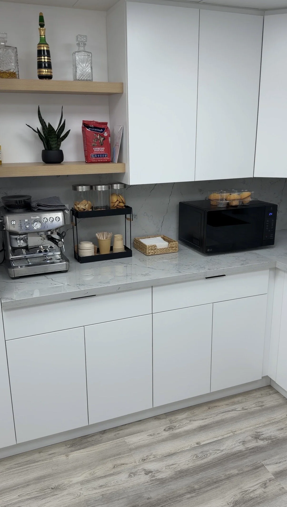 A modern kitchen countertop with a coffee machine, a microwave, a basket of snacks, and various containers. White cabinets and wood flooring are also visible.