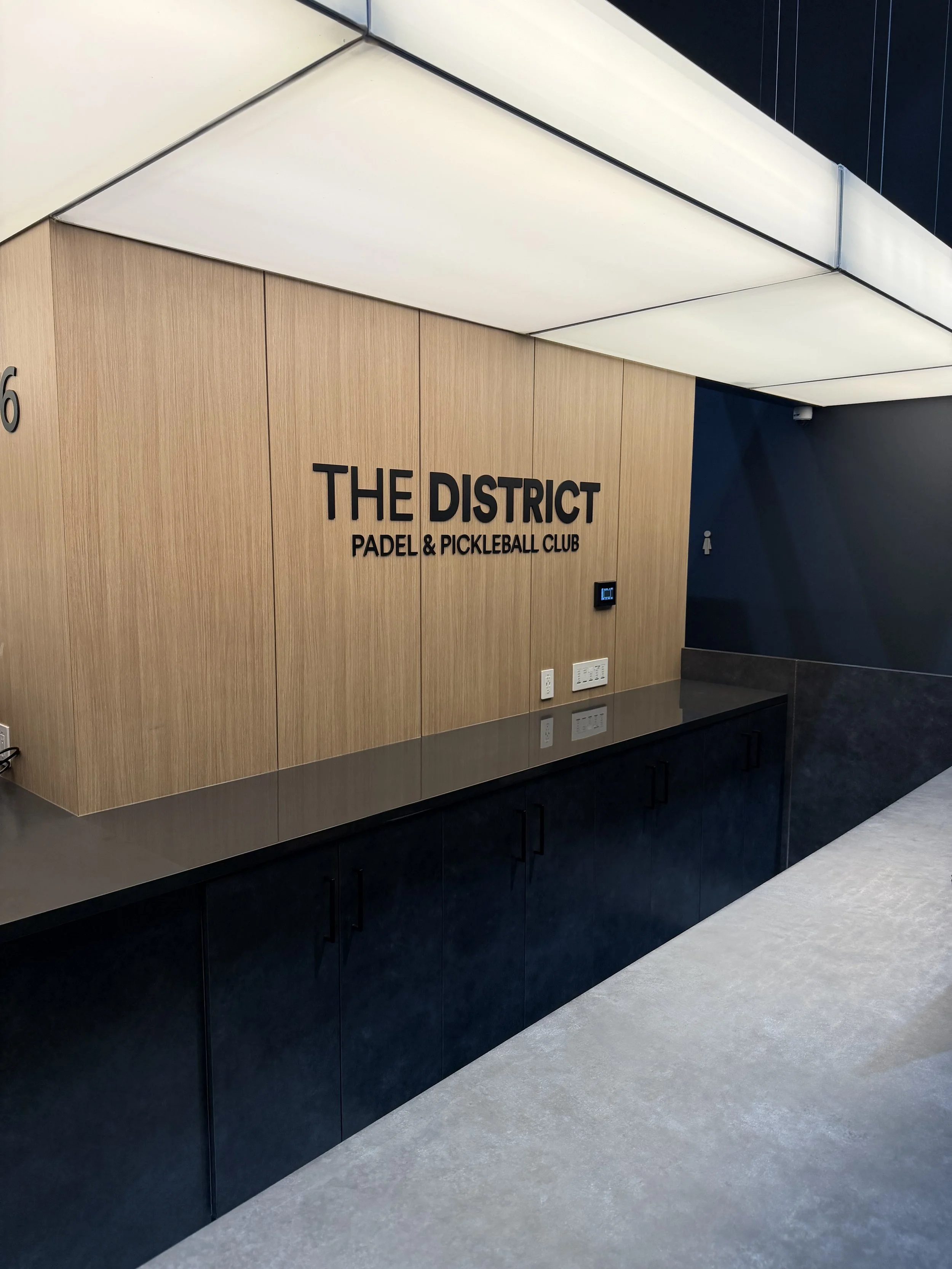 Wood-paneled wall with black text reading 'The District Padel & Pickleball Club', and black cabinetry below, in a modern interior space.